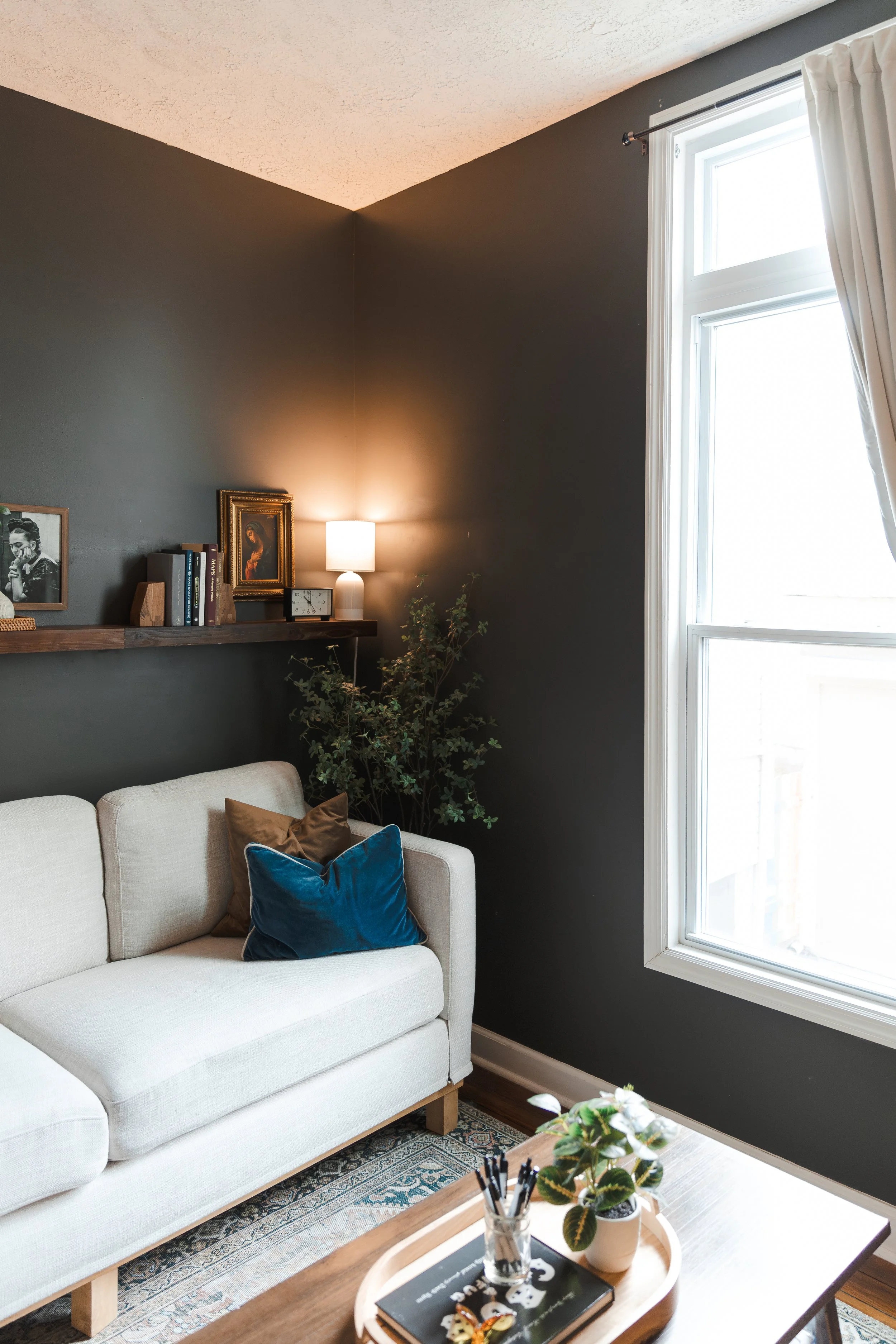 A cozy living room corner featuring a white sofa with brown and blue pillows, a wooden coffee table with plants and stationery, a dark accent wall with a floating shelf holding books and decor, a small lamp, and large windows with white curtains lett