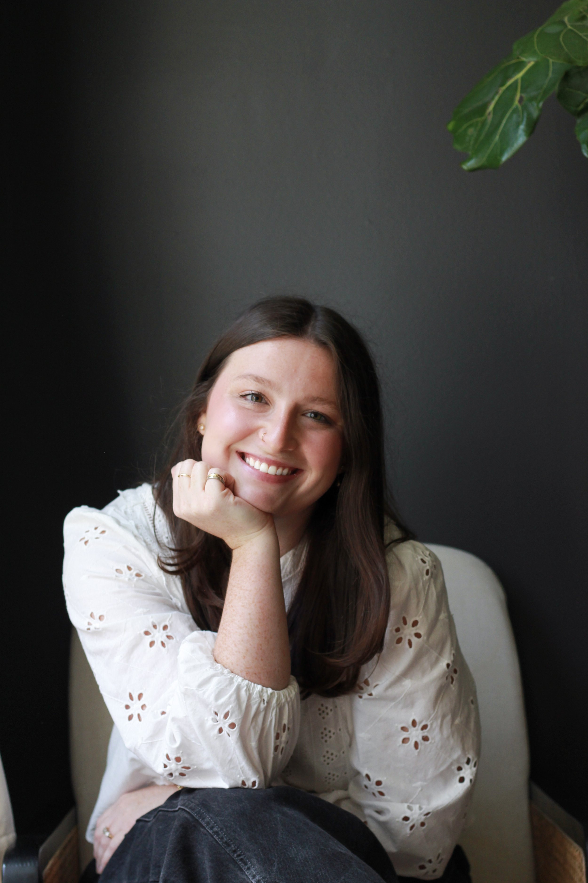 A woman with long brown hair, smiling, resting her chin on her right hand, wearing a white eyelet blouse and sitting in a light-colored chair against a dark wall with a green leafy plant in the upper right corner.