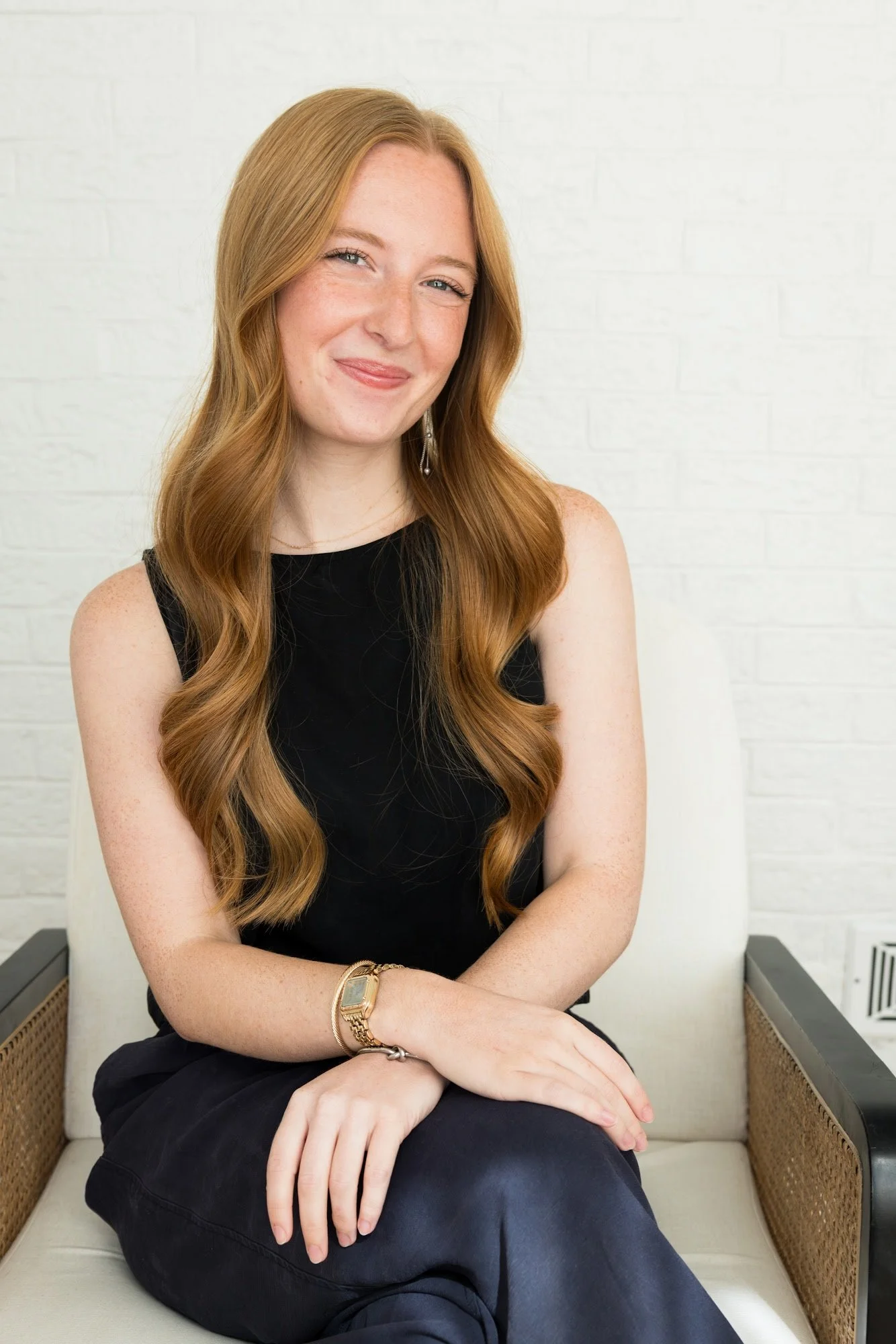 A woman with long, wavy auburn hair, wearing a white t-shirt and a gold watch, sitting on a beige chair against a dark gray wall, smiling softly.