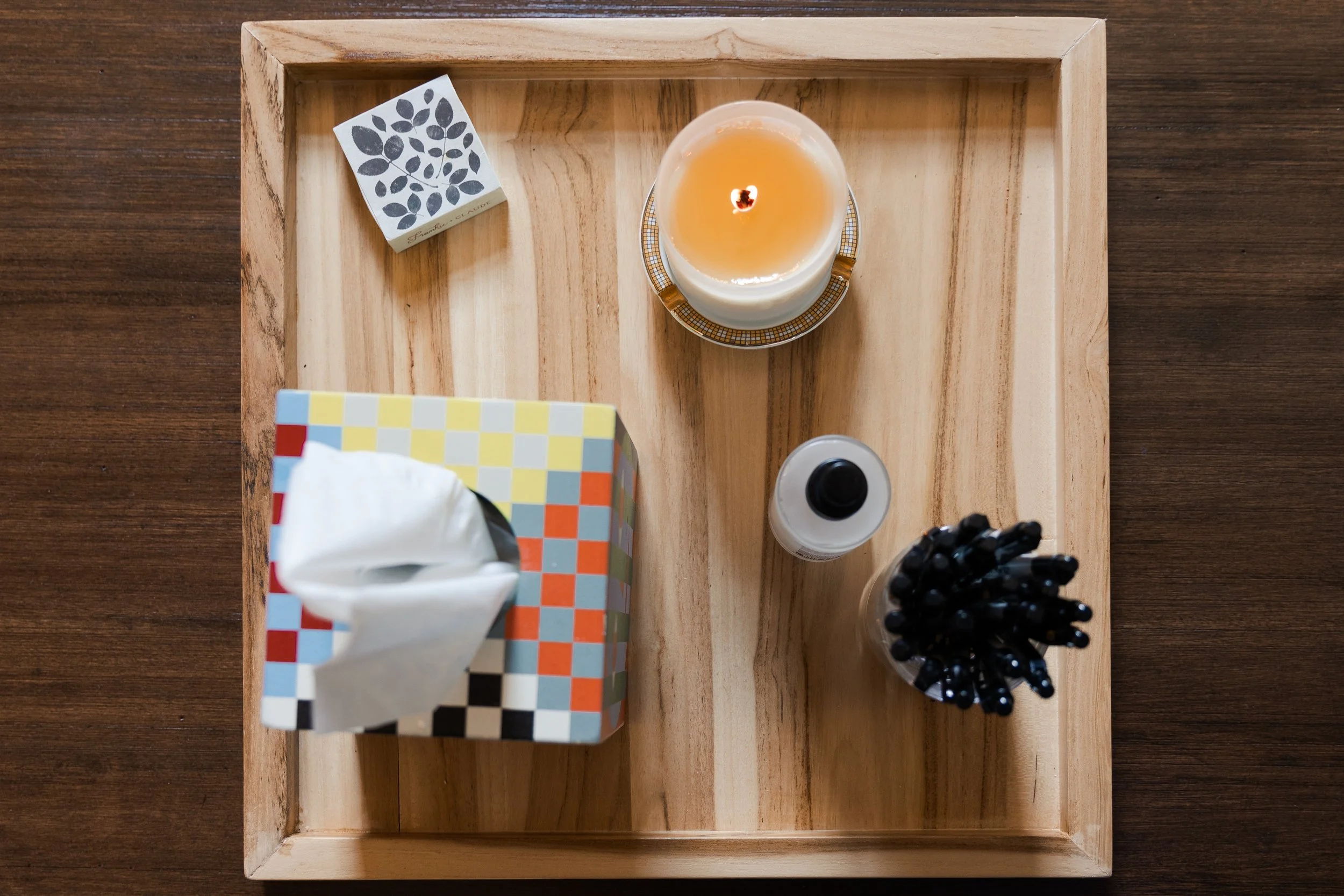 Square wooden tray with a candle, tissue box, small bottle, and black pens on a wooden table.