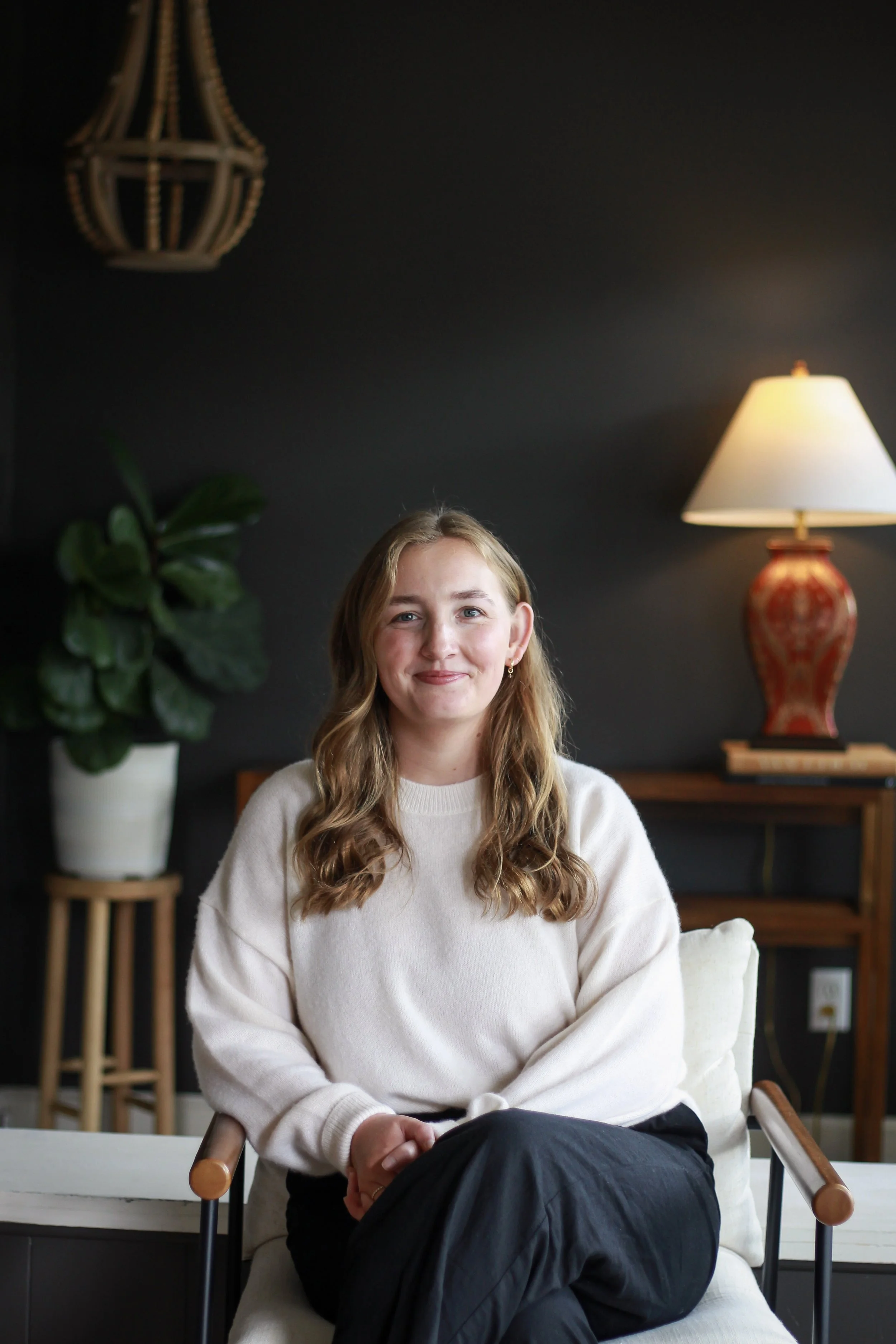 A young woman with light brown hair, sitting in a chair with a white sweater, smiling at the camera. Behind her is a dark wall, a potted plant, a table with a lamp, and a wood shelf.