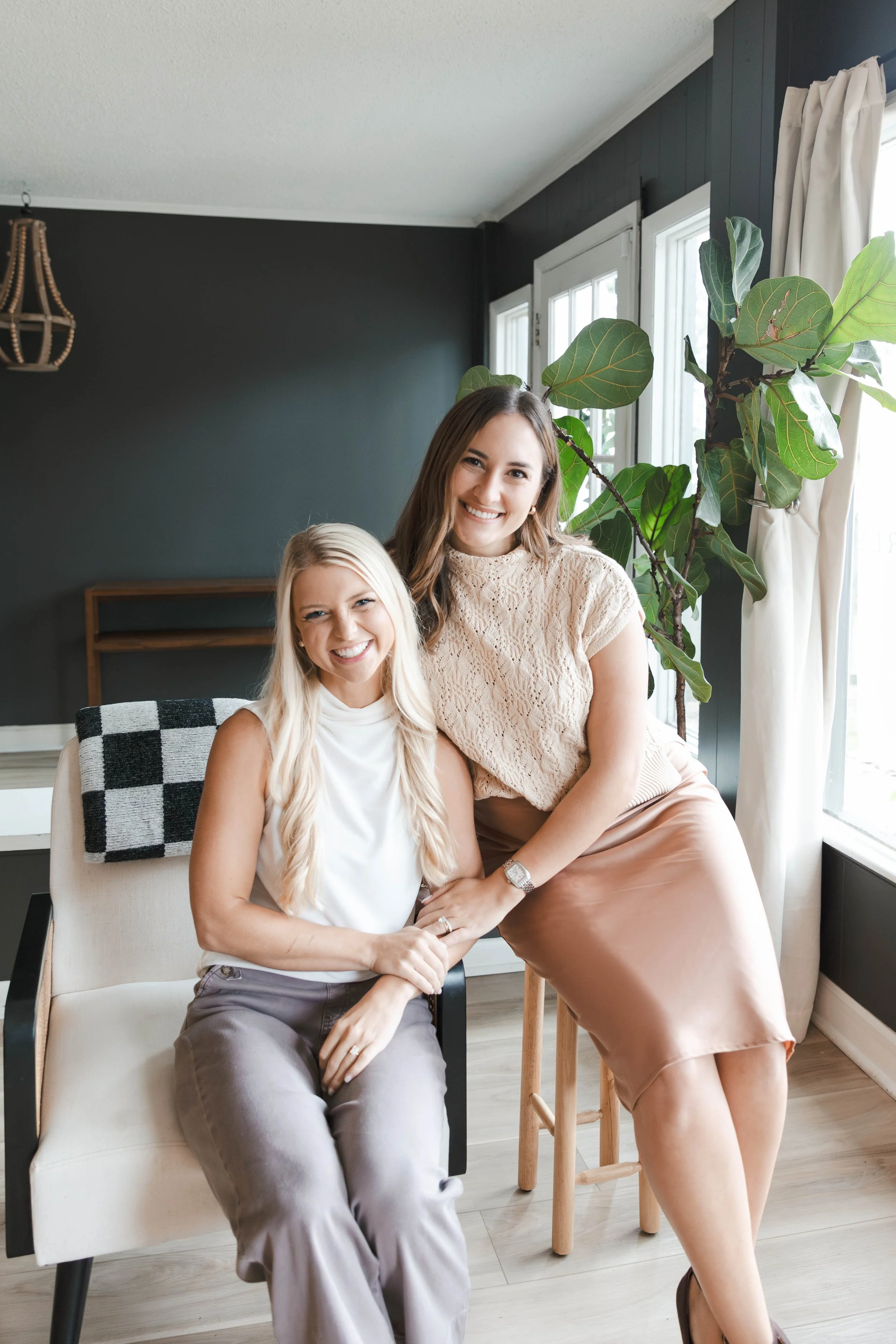 Two smiling women, one with long blonde hair sitting on a chair and the other with long brown hair standing behind her, indoors near a large window with a green plant and dark wall in the background.