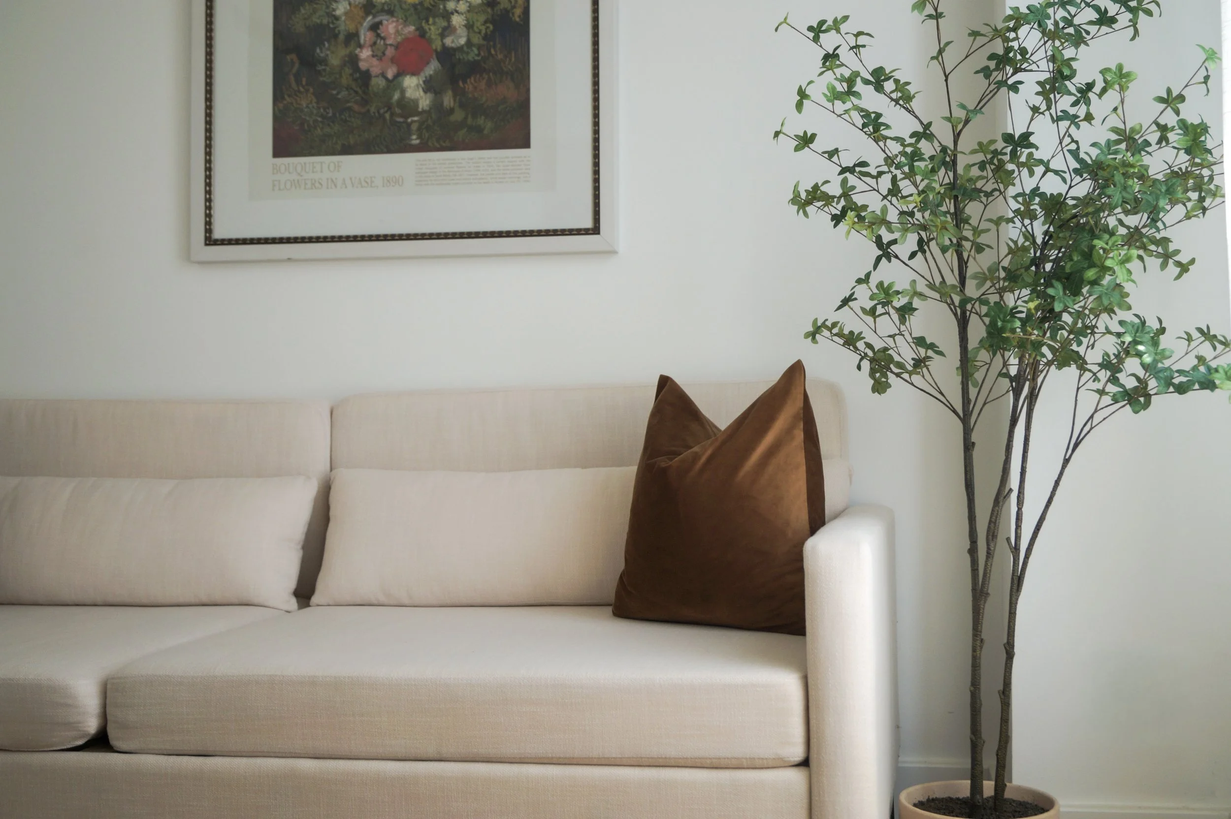 A beige sofa with a brown pillow on the right side, next to a tall potted green plant, with a framed floral artwork hanging on the white wall above the sofa.