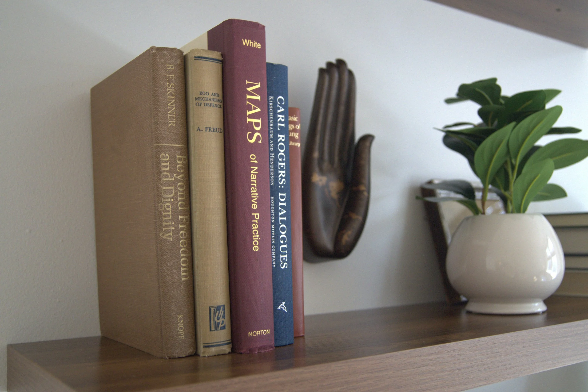 A wooden bookshelf with five books and a decorative hand sculpture, with a white ceramic pot with green leaves and a closed book in the background.