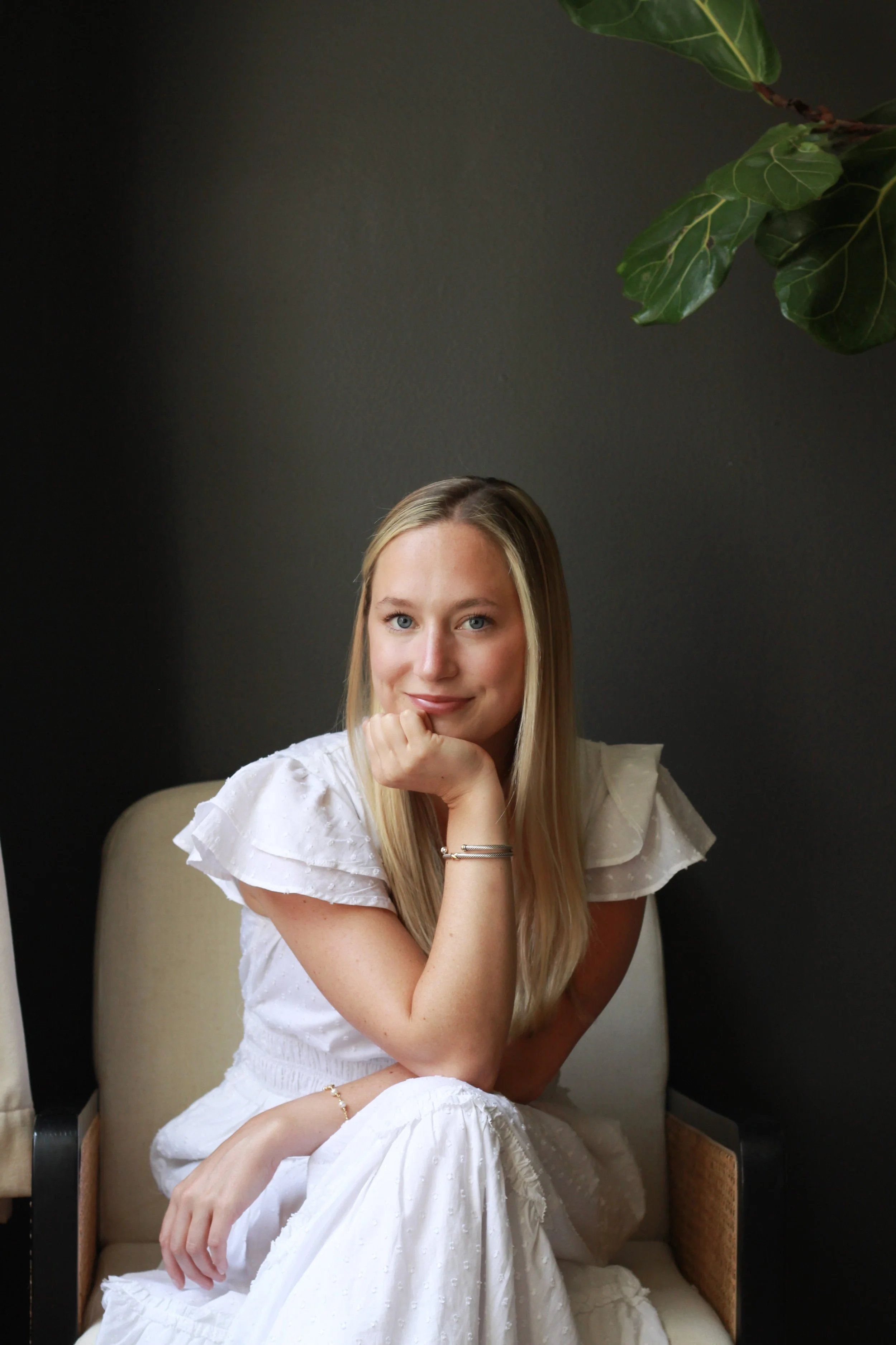 A young woman with long blonde hair, wearing a white dress with ruffled sleeves, smiling and resting her chin on her hand, sitting on a beige chair against a dark wall, with a leafy green plant visible in the upper right corner.
