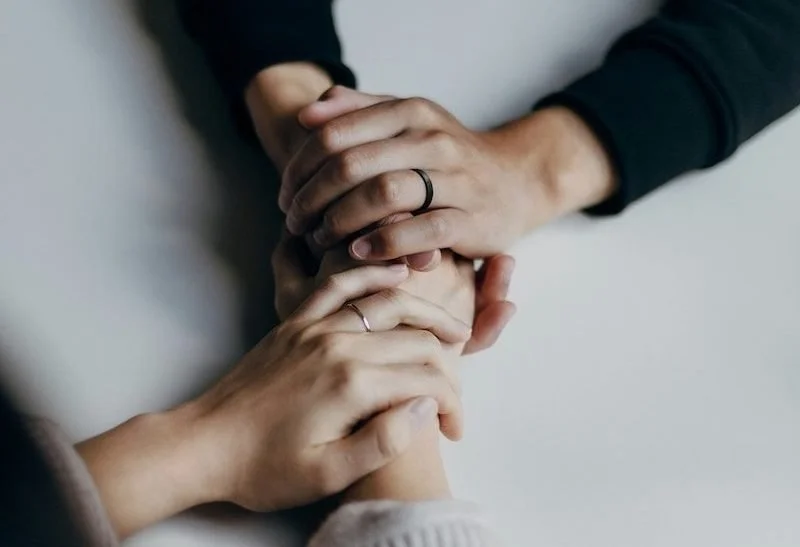 Two people holding hands, one with a wedding ring, on a white surface.