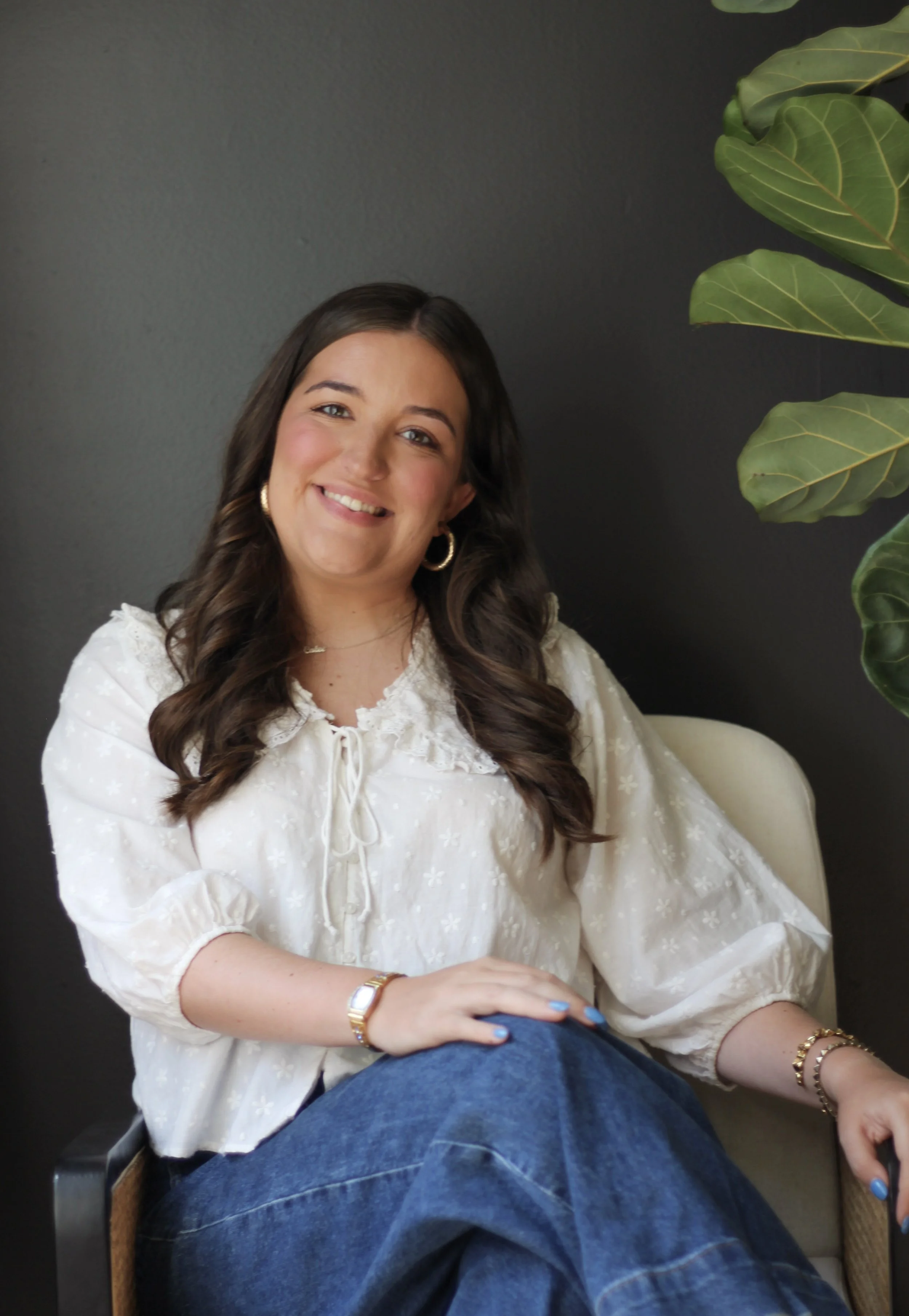 A young woman with long brown hair, wearing a white blouse with embroidered details, gold hoop earrings, a watch, and bracelets, sitting on a chair against a dark gray wall and a large green houseplant.