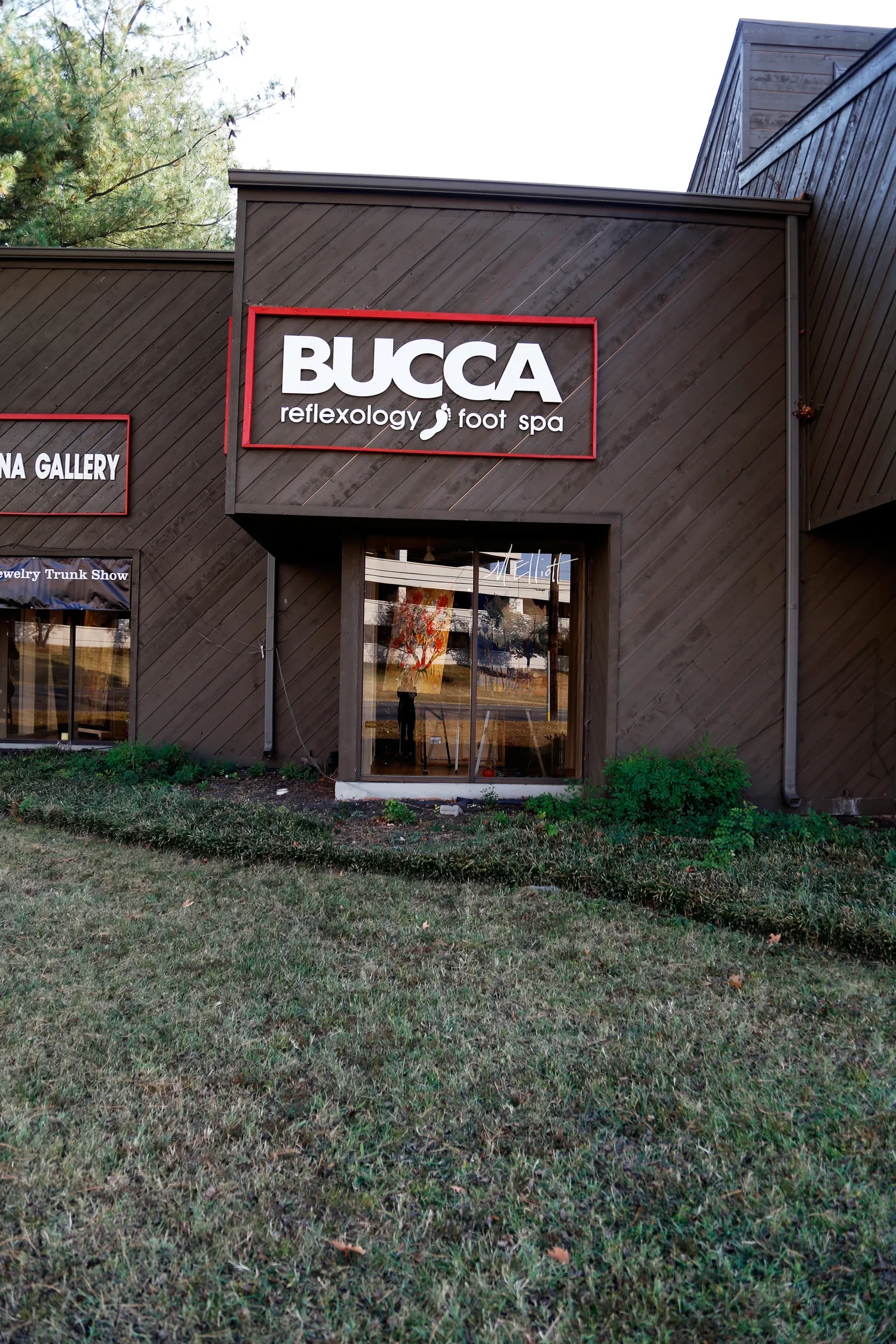 Exterior view of a building with a sign that reads 'BUCCA reflexology foot spa', featuring dark wooden siding and a large window reflecting colorful artwork.