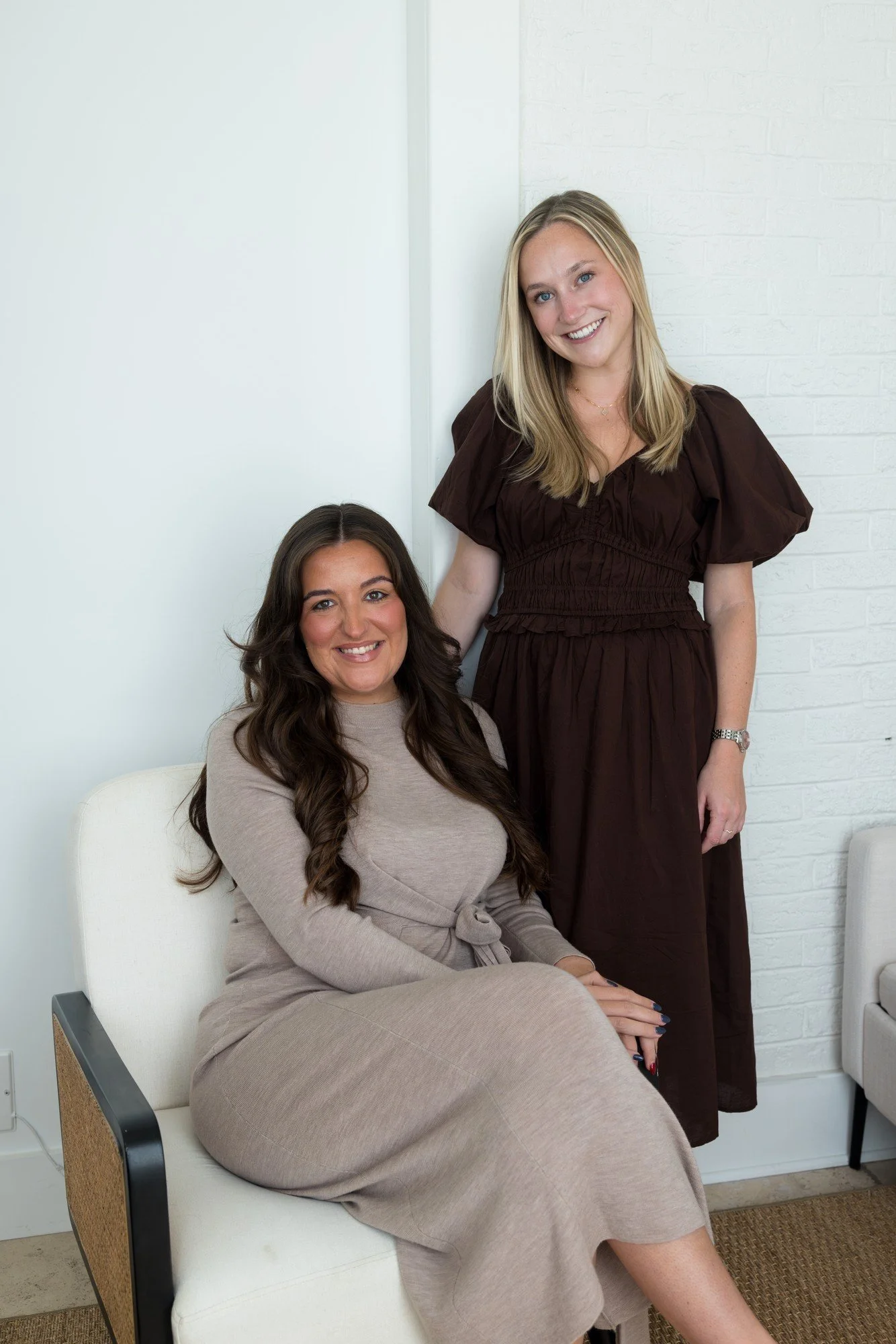 Two women smiling, one sitting on a white chair and the other standing beside her, in a room with white walls.