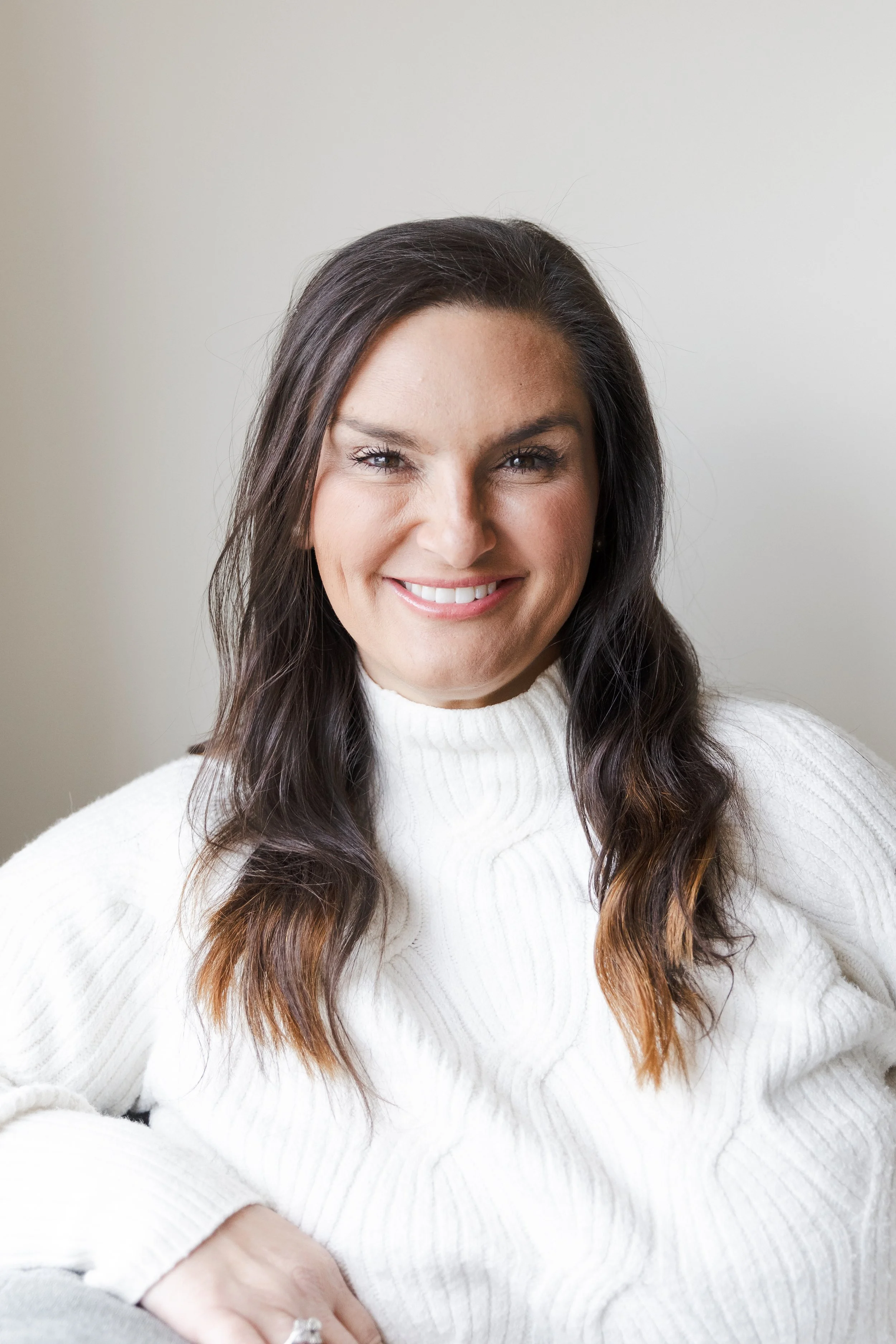 Close-up of a smiling woman with dark brown hair and light skin, wearing a white sweater, sitting against a plain light background.