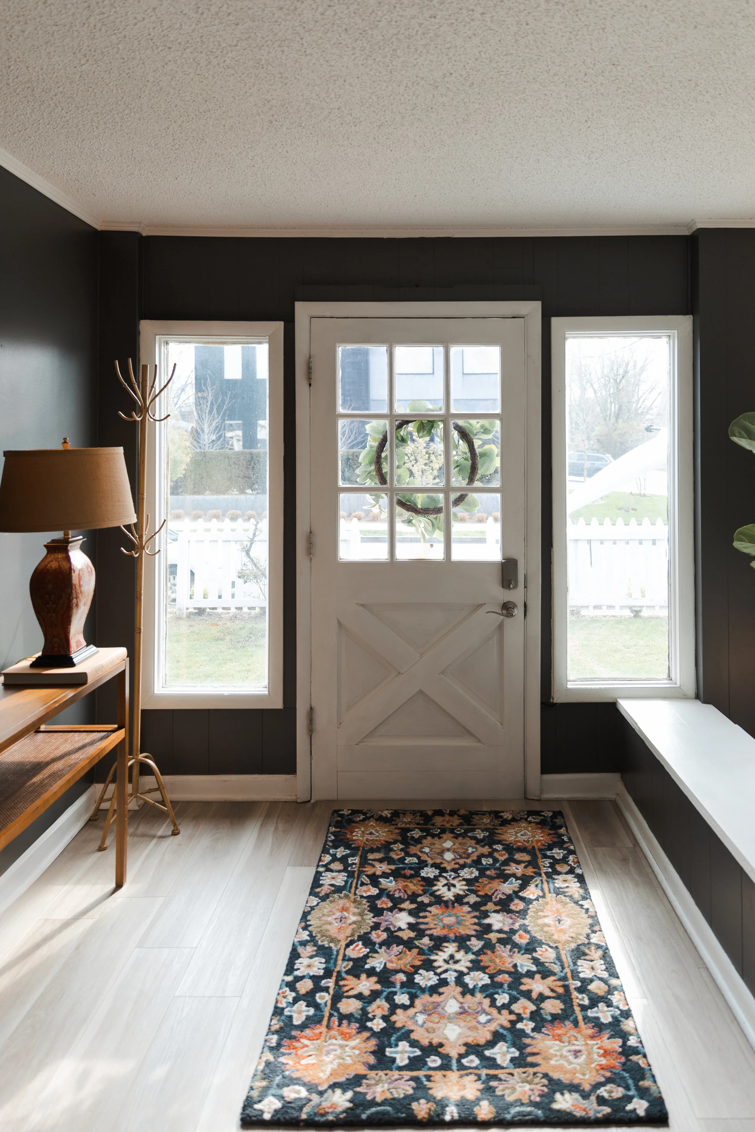 Front door with a wreath, flanked by two narrow windows, and a colorful floral rug on light-colored floor in a home entryway.