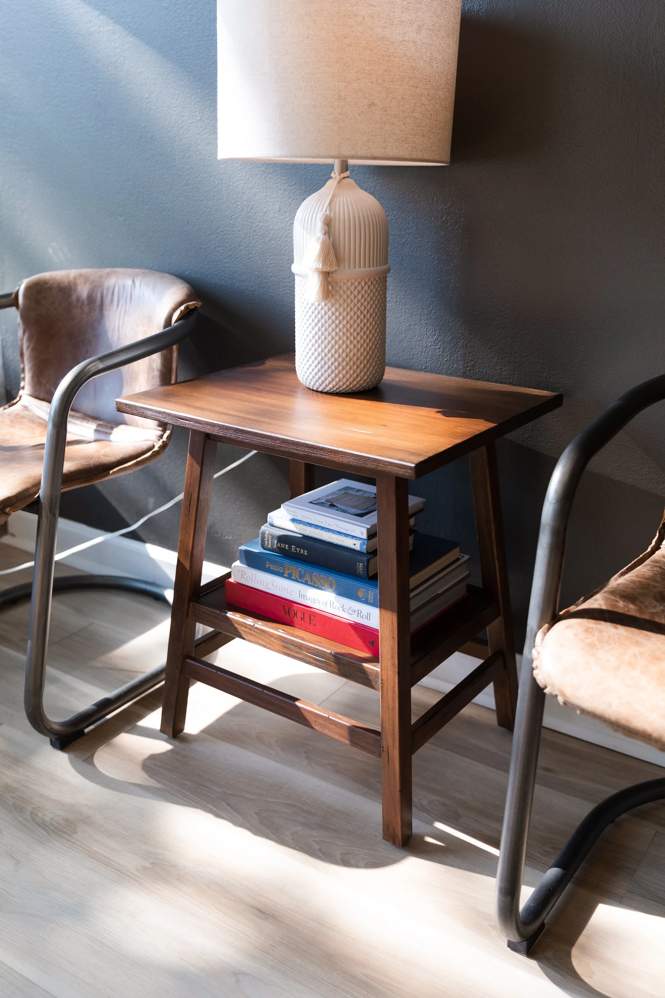 A wooden side table with a lamp on top, surrounded by two chairs with worn leather seats, and a stack of books underneath.