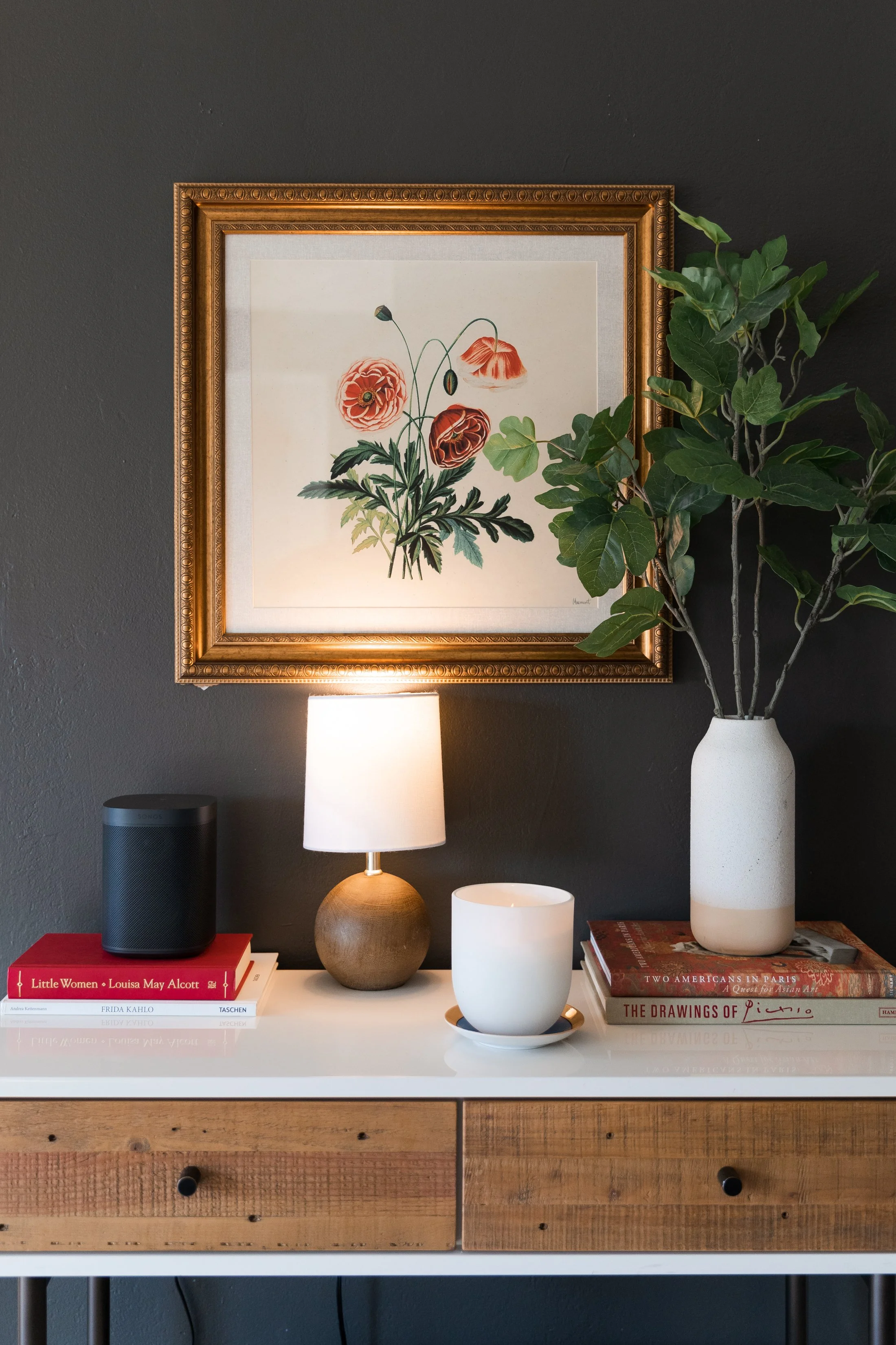 A home decor arrangement on a white and wood sideboard against a dark gray wall, featuring a framed floral print, a small table lamp, a ceramic cup with a matching saucer, a potted plant in a tall vase, and several books.