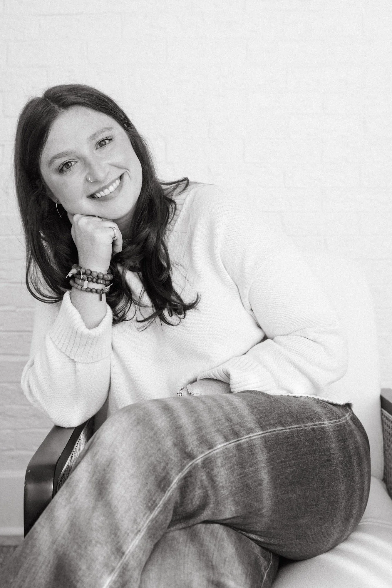 A woman with long brown hair, smiling, sitting on a beige chair against a dark wall, resting her chin on her hand, wearing a white eyelet blouse and black jeans.