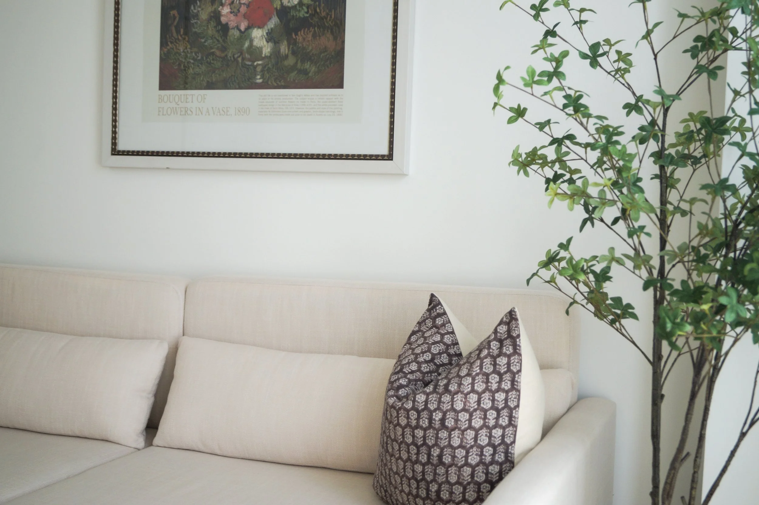 A white sofa with two dark-patterned pillows, a large window with a framed picture titled 'Bouquet of Flowers in a Vase, 1890' above it, and a tall green plant on the right side.