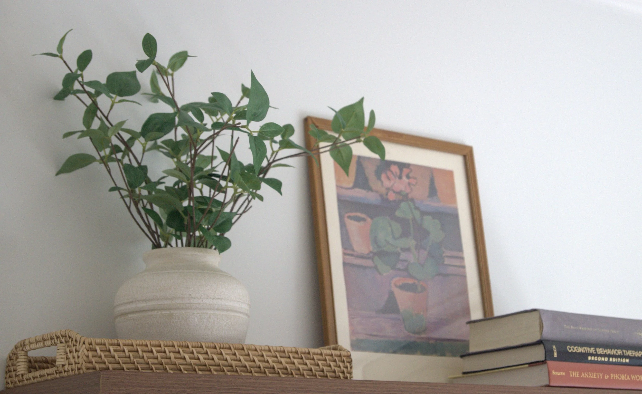Interior scene with a potted green plant, framed painting of potted plants, and stacked books on a wooden surface.