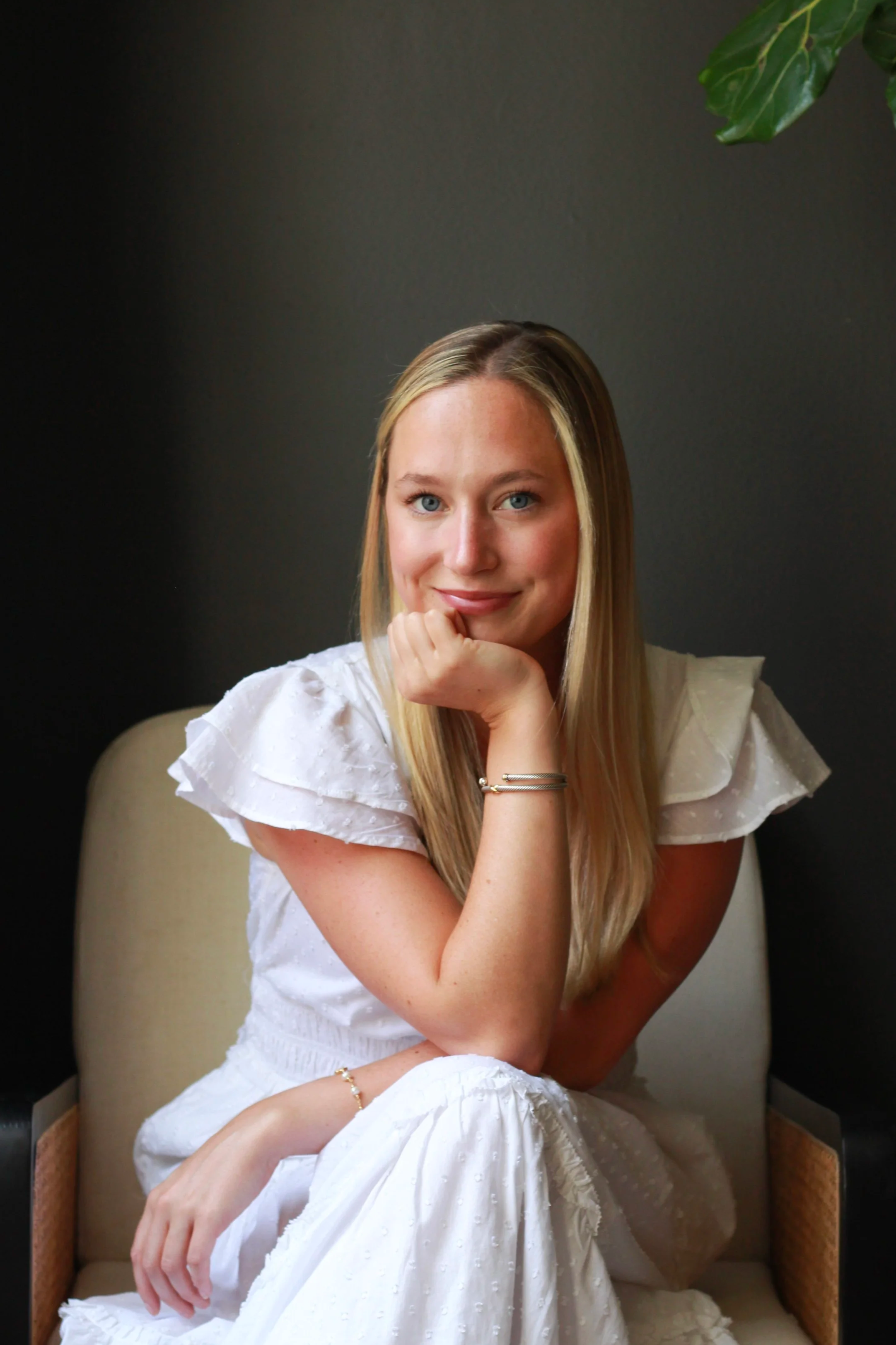 A young woman with blonde hair and blue eyes is sitting on a beige chair against a dark wall, smiling and resting her chin on her hand, wearing a white dress with ruffled sleeves and jewelry.