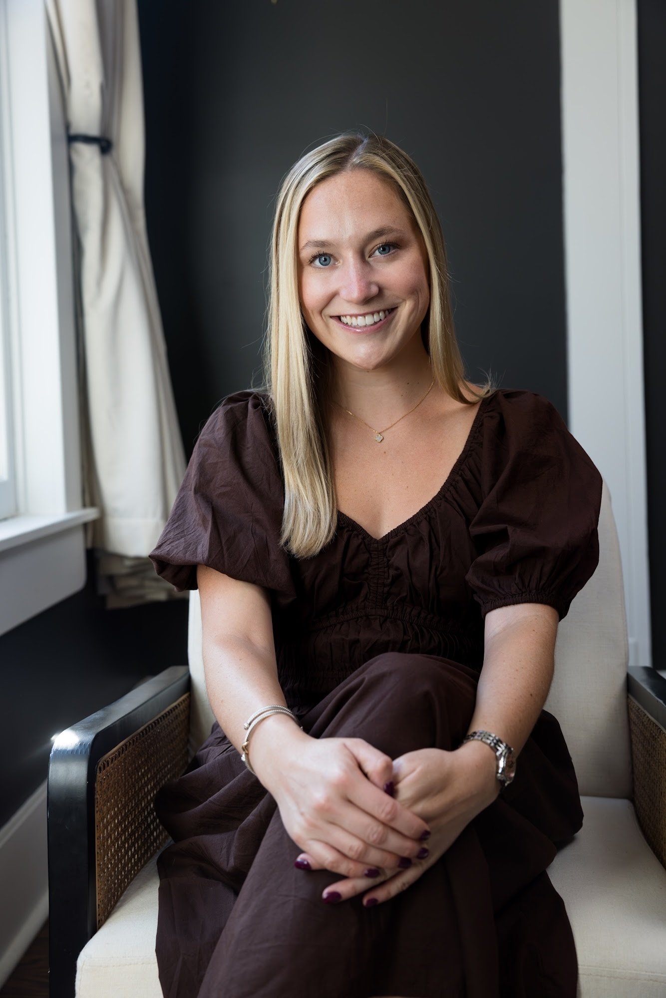 A smiling woman with blonde hair sitting on a beige armchair against a dark gray wall, wearing a white dress with ruffled sleeves.
