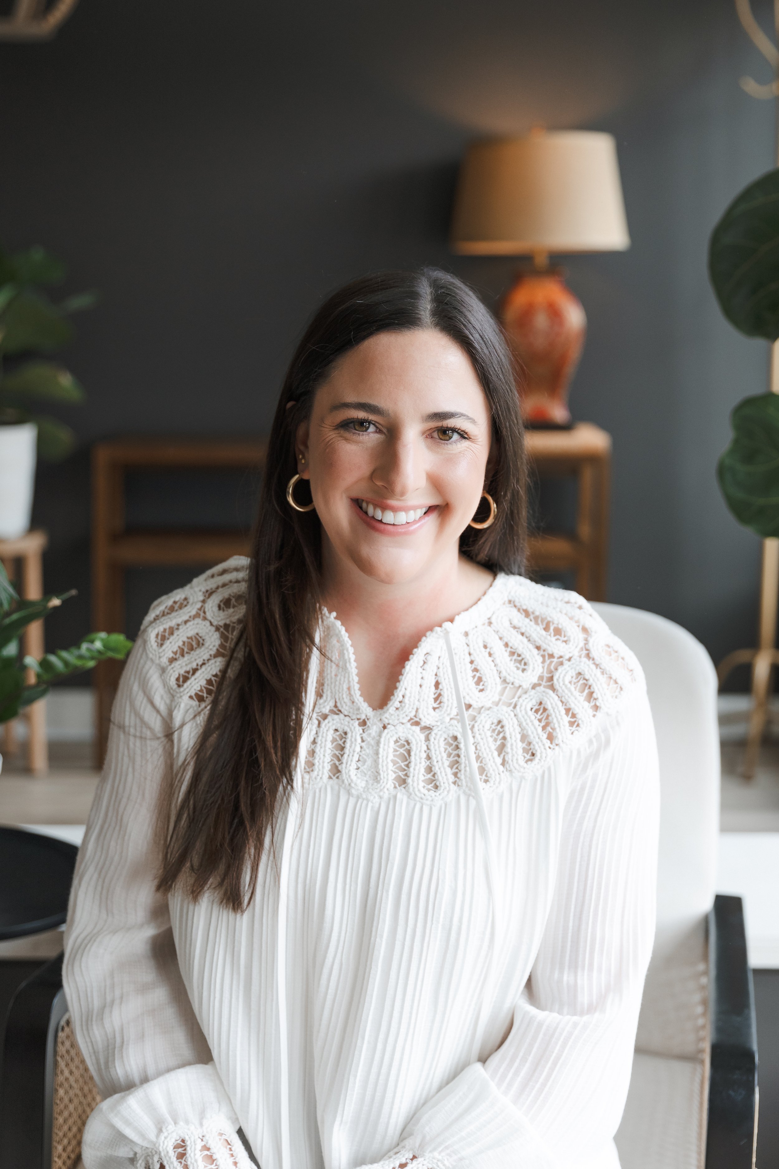 A woman with long dark hair, wearing a white lace blouse and gold hoop earrings, sitting in a modern living room with plants and a table with a lamp in the background.