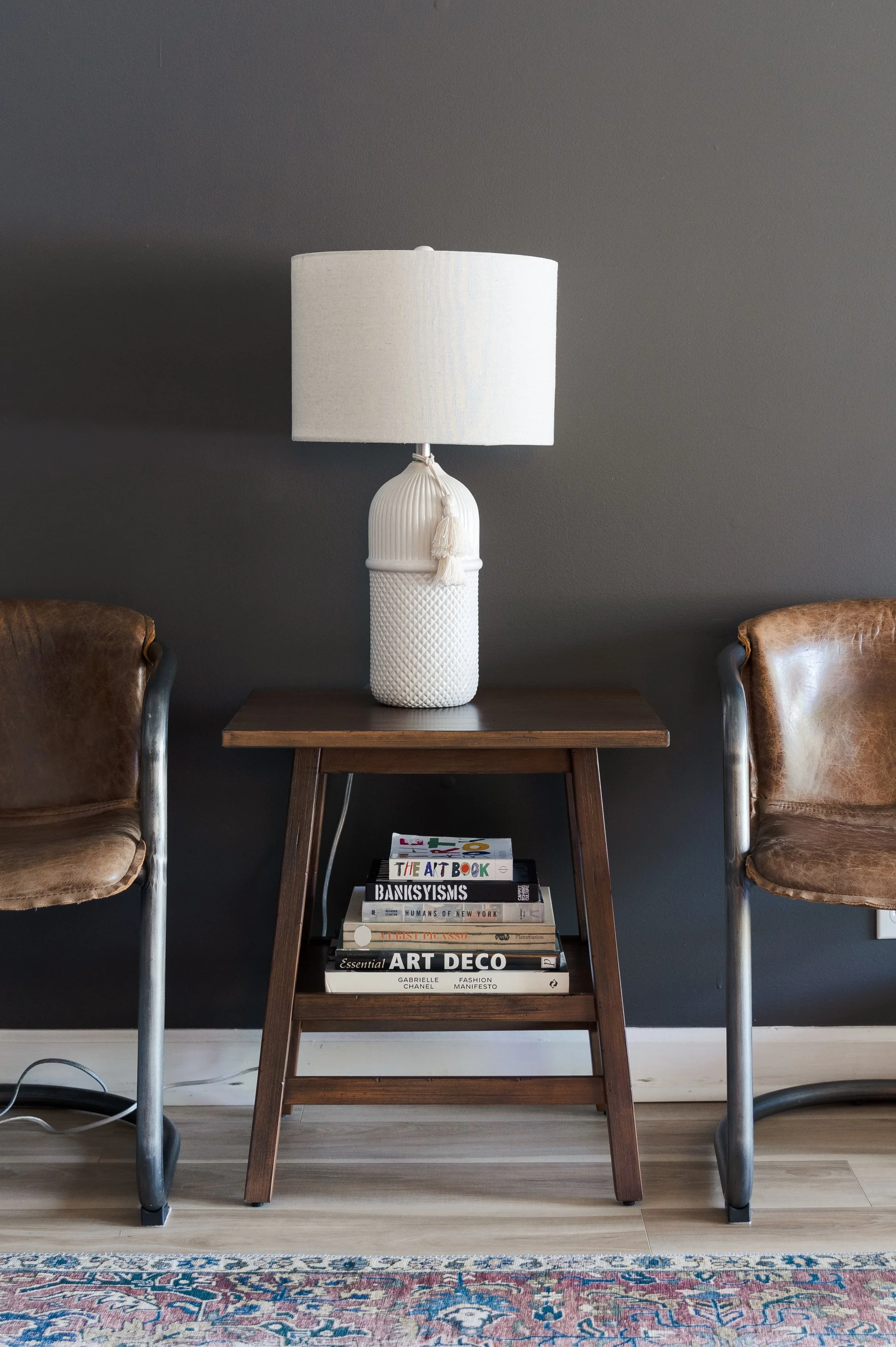 A living room with two brown leather armchairs on either side of a small wooden side table. The table holds a tall white ceramic lamp with a large drum shade and a tassel decoration. A stack of books is on a lower shelf of the table. The background wall is painted dark gray, and the floor has a light-colored wood finish with a patterned area rug.