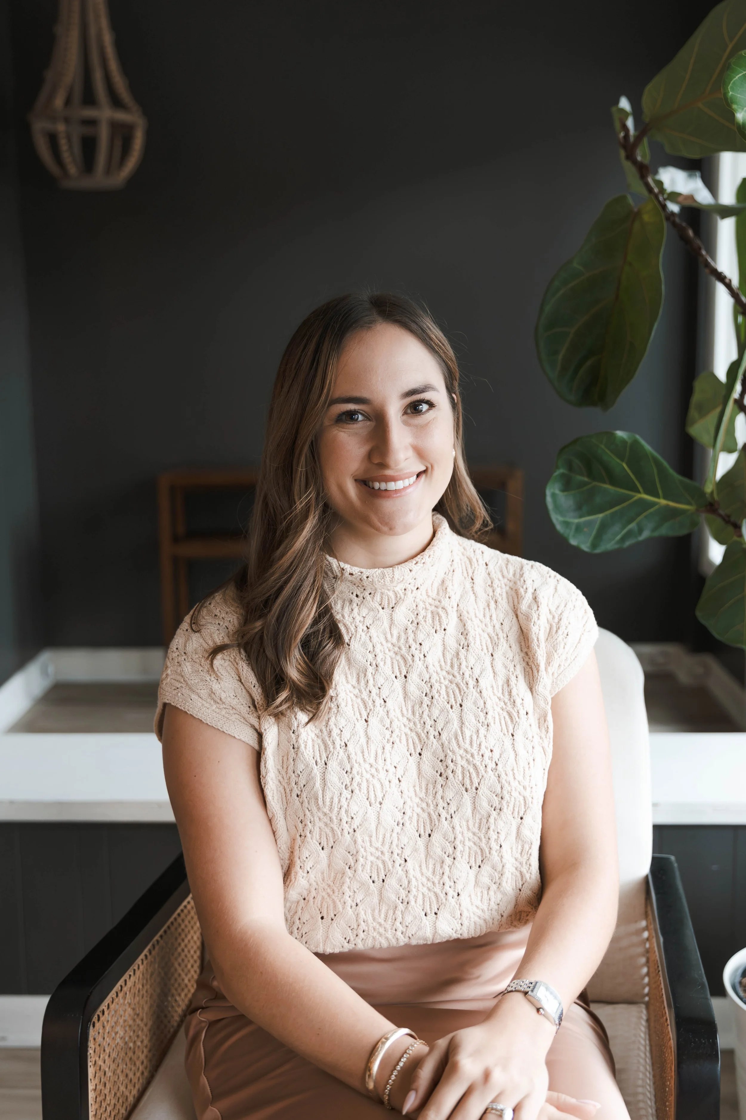 A young woman with long brown hair smiling and sitting in a room with dark walls and natural light from a window, wearing a cream-colored lace top and a watch.