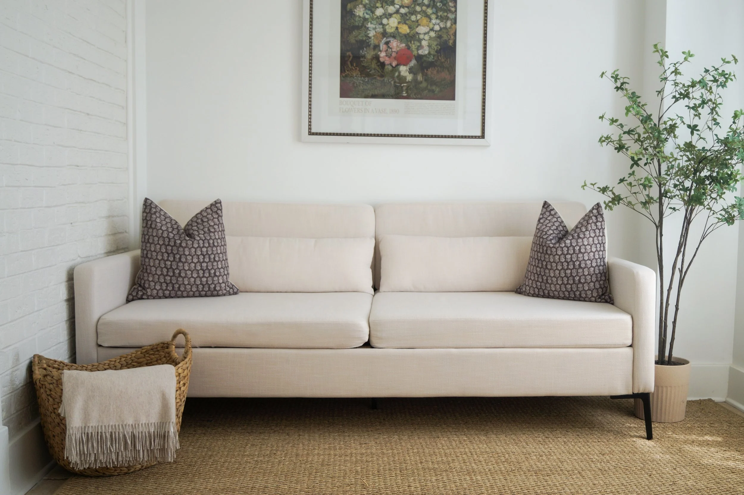 A cream-colored sofa with two patterned throw pillows in a living room with a framed floral artwork on the white wall, a potted plant, and a basket with a blanket.