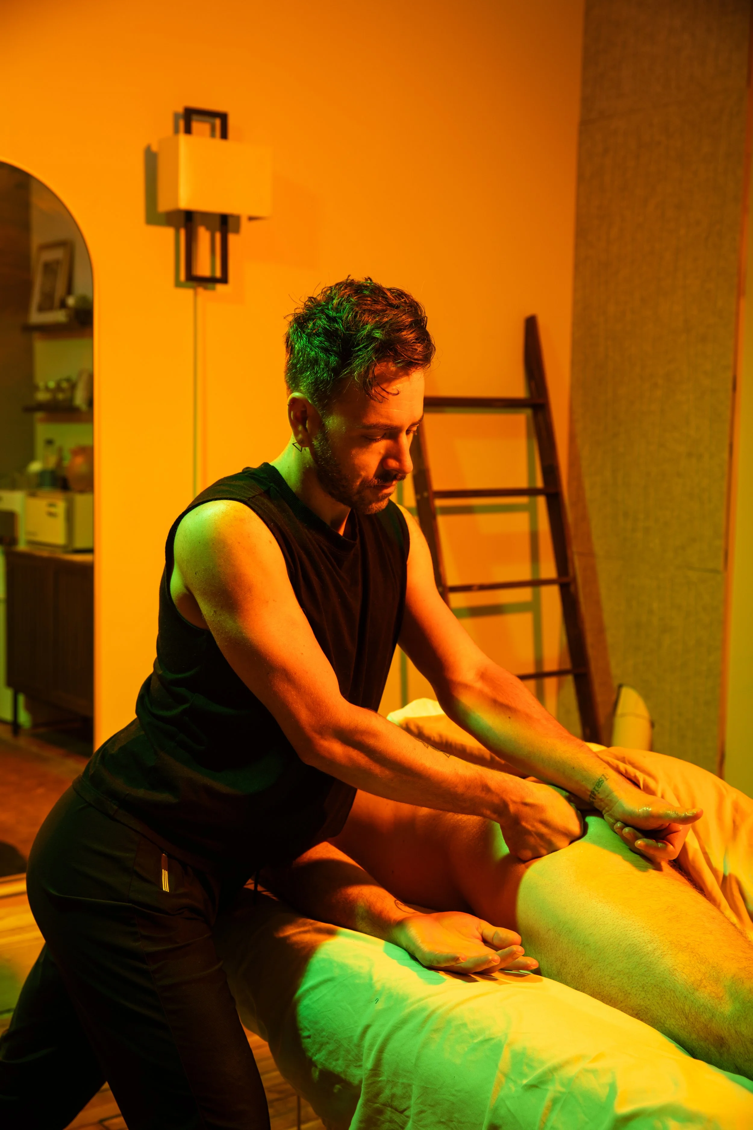 A man giving a massage to a person lying face down on a massage table, with warm orange lighting in the room.