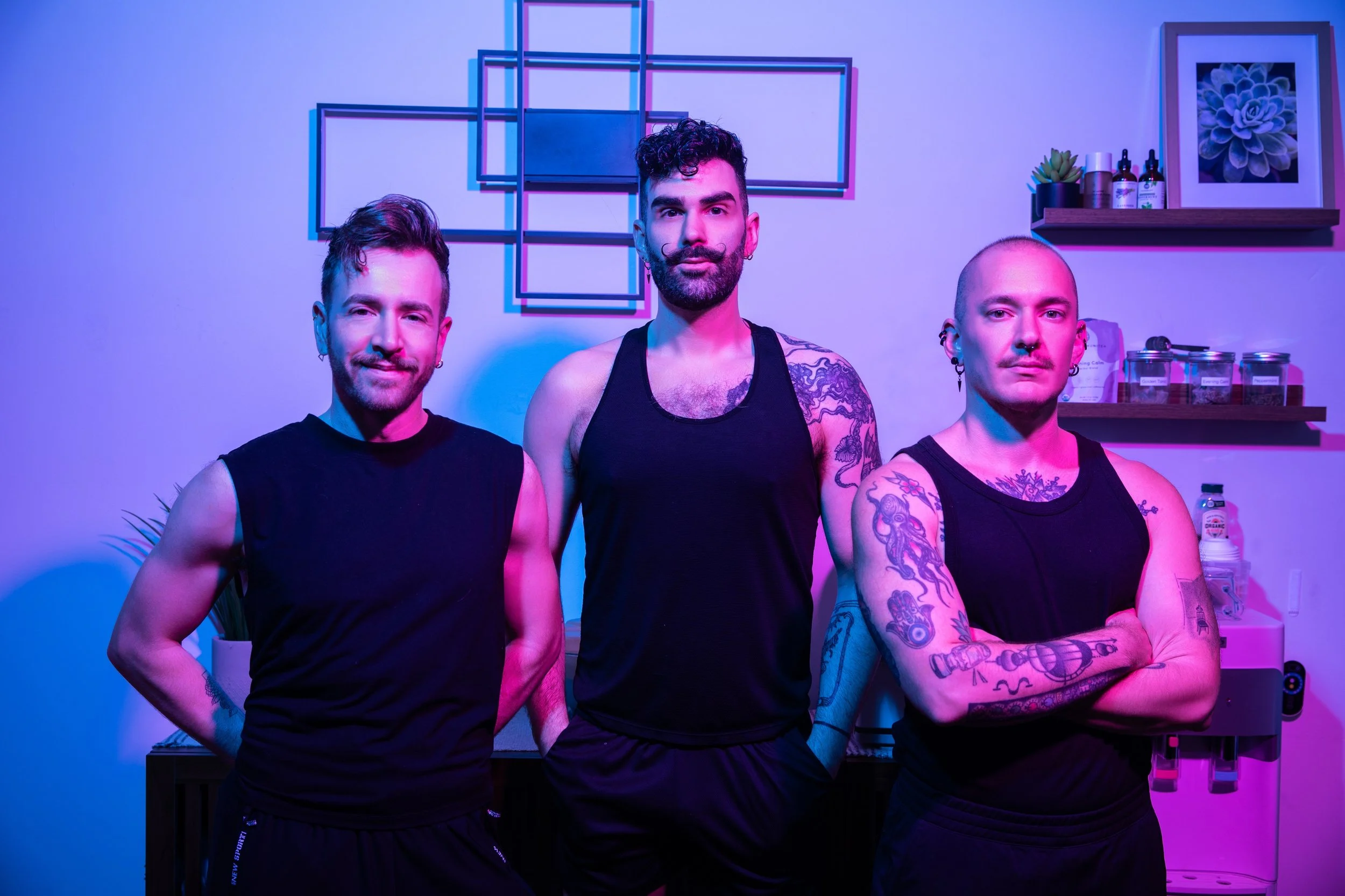 Three men standing together in a room with purple and blue lighting. They are dressed in black tank tops and have various tattoos and facial piercings. Behind them are shelving units with plants, bottles, and jars.