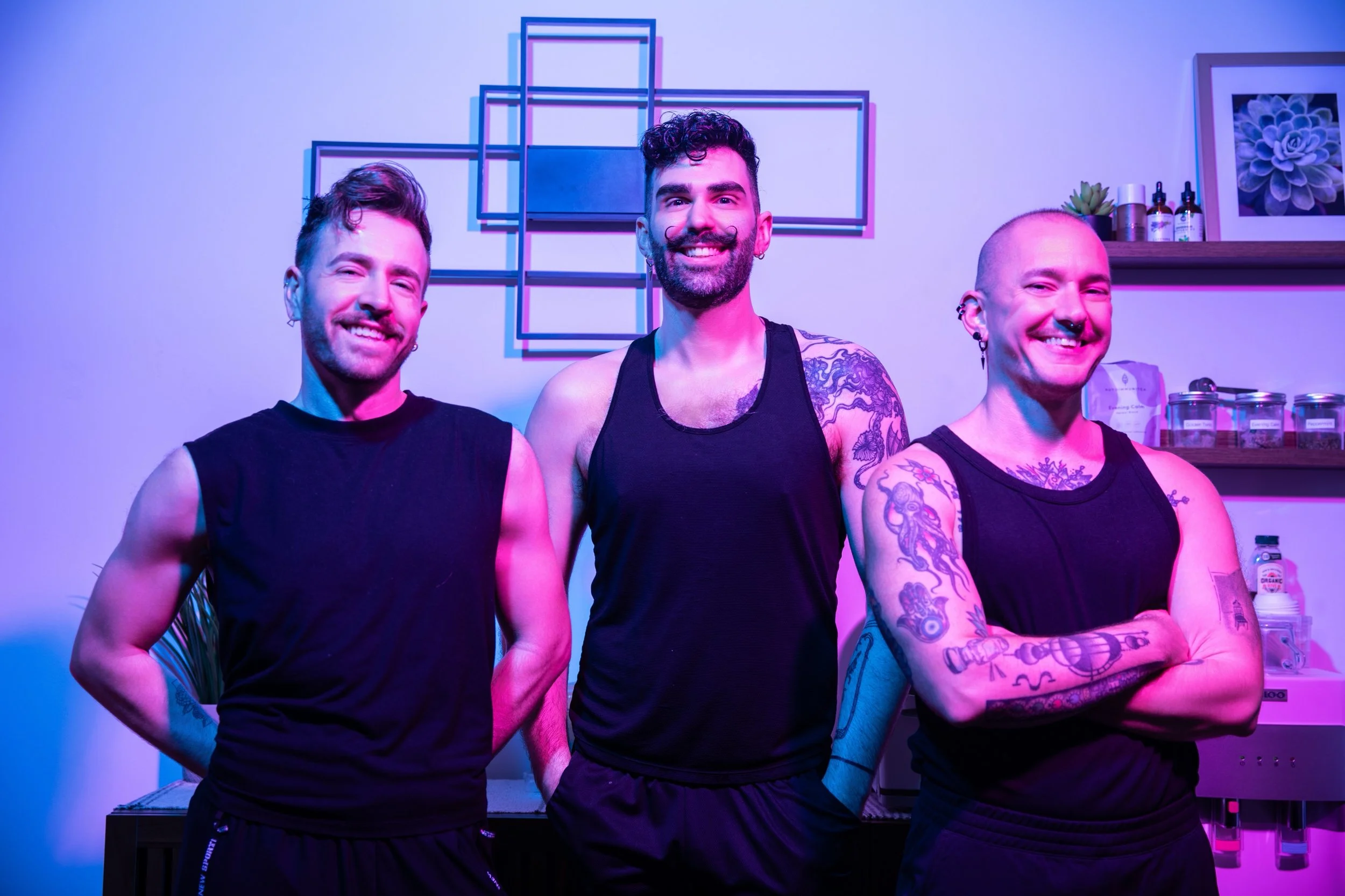 Three smiling men in black tank tops standing together in a room with purple and blue lighting. The background features abstract wall art and shelves with bottles and plants.