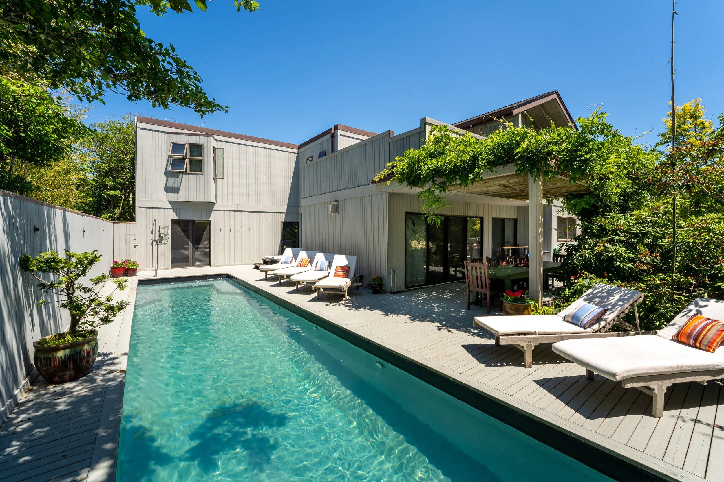 A backyard with a swimming pool, white lounge chairs with colorful pillows, potted plants, and a house with a deck, surrounded by green trees under a clear blue sky.