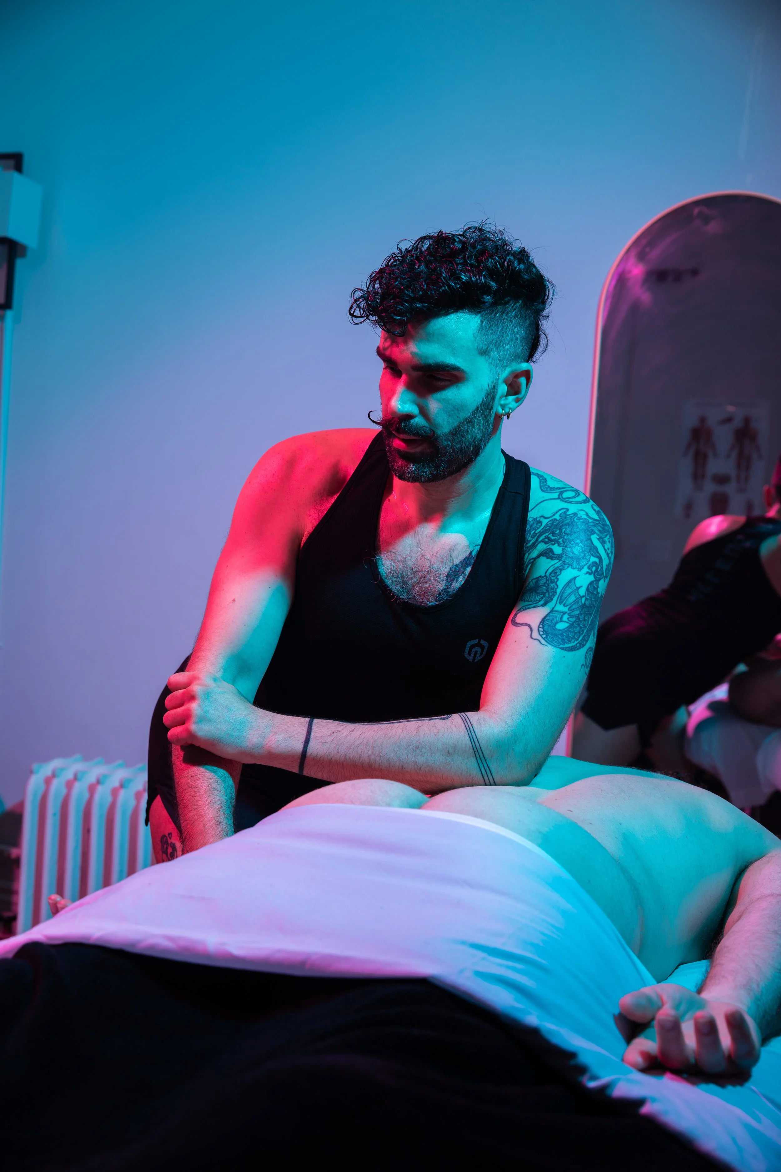 A man with tattoos and a beard, wearing a black tank top, leaning over a person lying down on an examination table, in a room with blue and pink lighting, with a mirror reflecting a restroom sign.