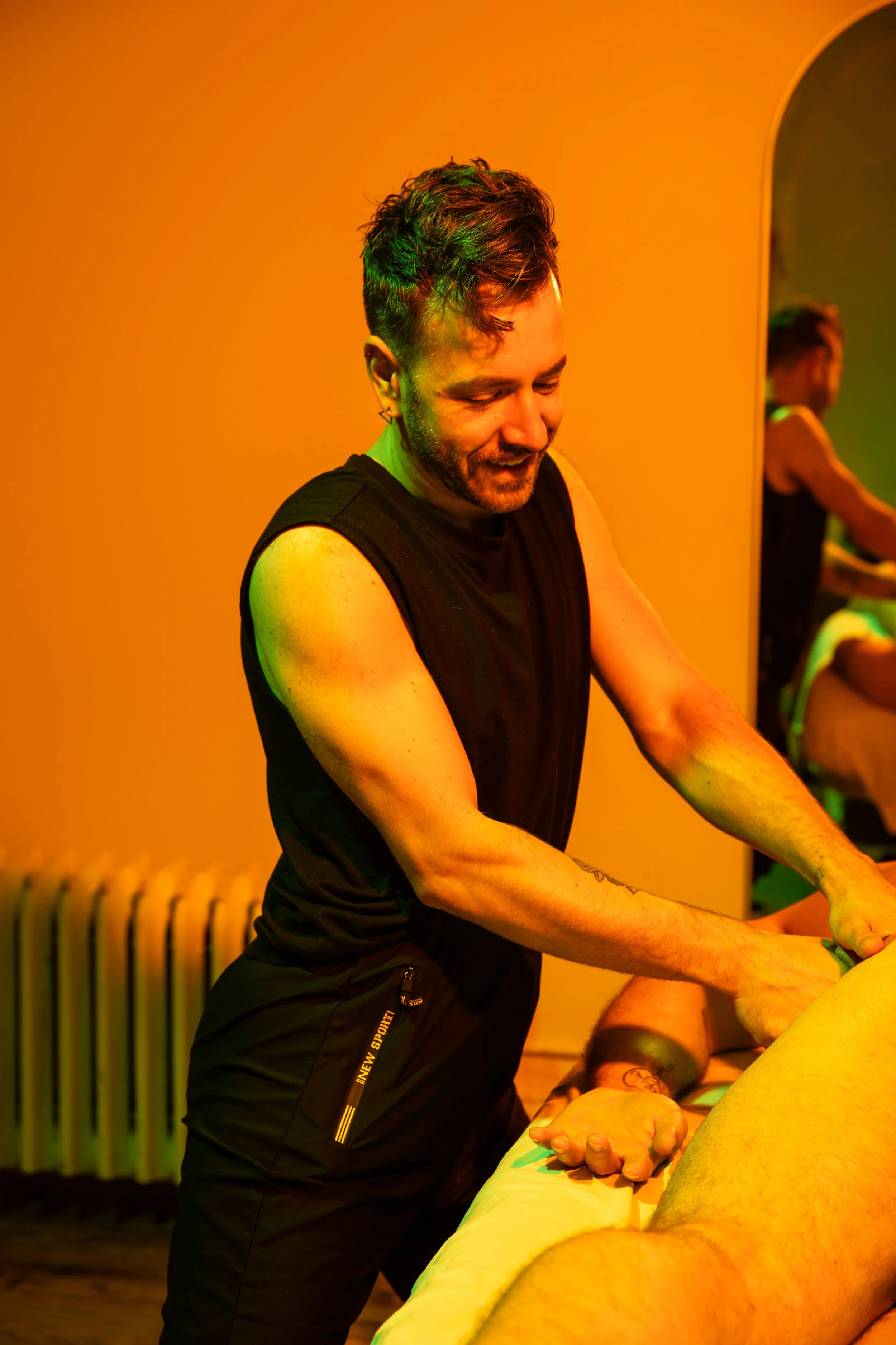 A man with short, curly hair and a beard, smiling, wearing a sleeveless black shirt, leaning over a massage table with a person receiving a massage, in a warmly lit room with orange walls and a mirror reflecting the scene.