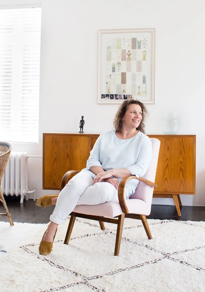 Une femme souriante assise sur une chaise rose dans un salon lumineux avec une armoire en bois derrière elle, une plante en verre, un cadre d'art moderne au mur, et un tapis blanc.