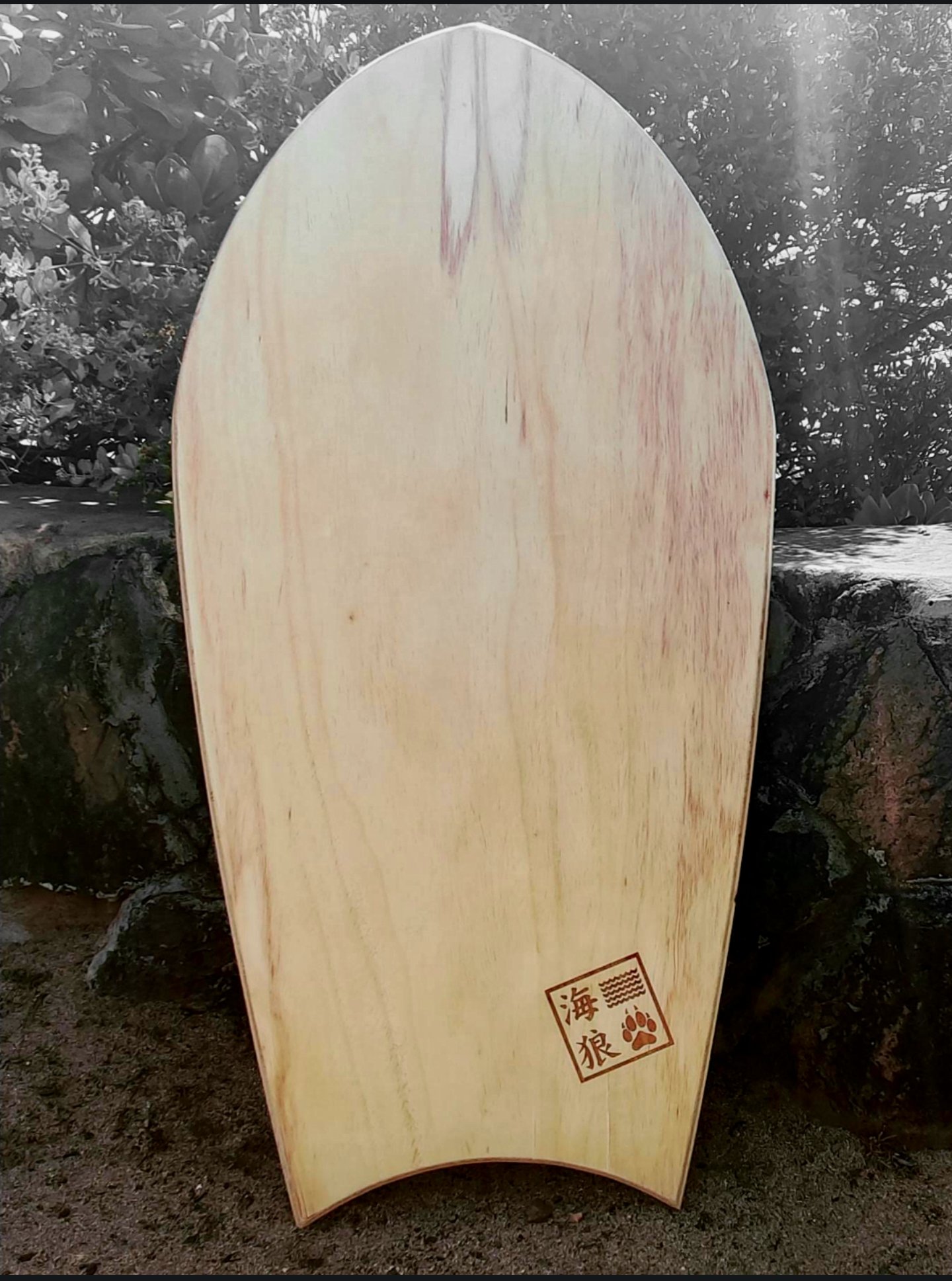 A pae po'o - Hawaiian wooden belly board diplayed upright, outdoors on sandy ground with a rock wall in the background.