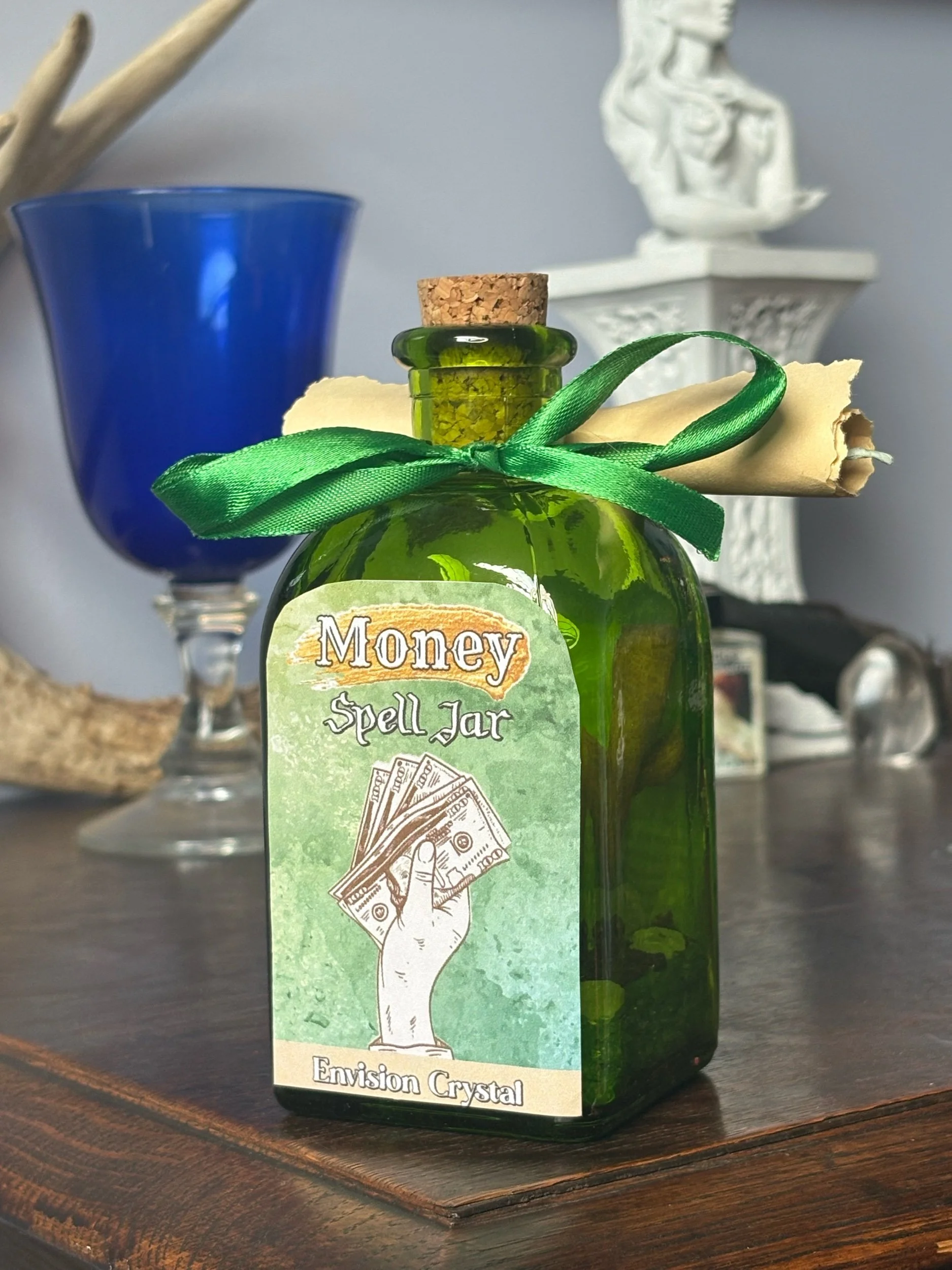 A green glass jar labeled 'Money Spell Jar' with a cork stopper, a rolled-up paper tied with a green ribbon, herbs inside. The jar is on a wooden surface with decorative items and a blue glass in the background.