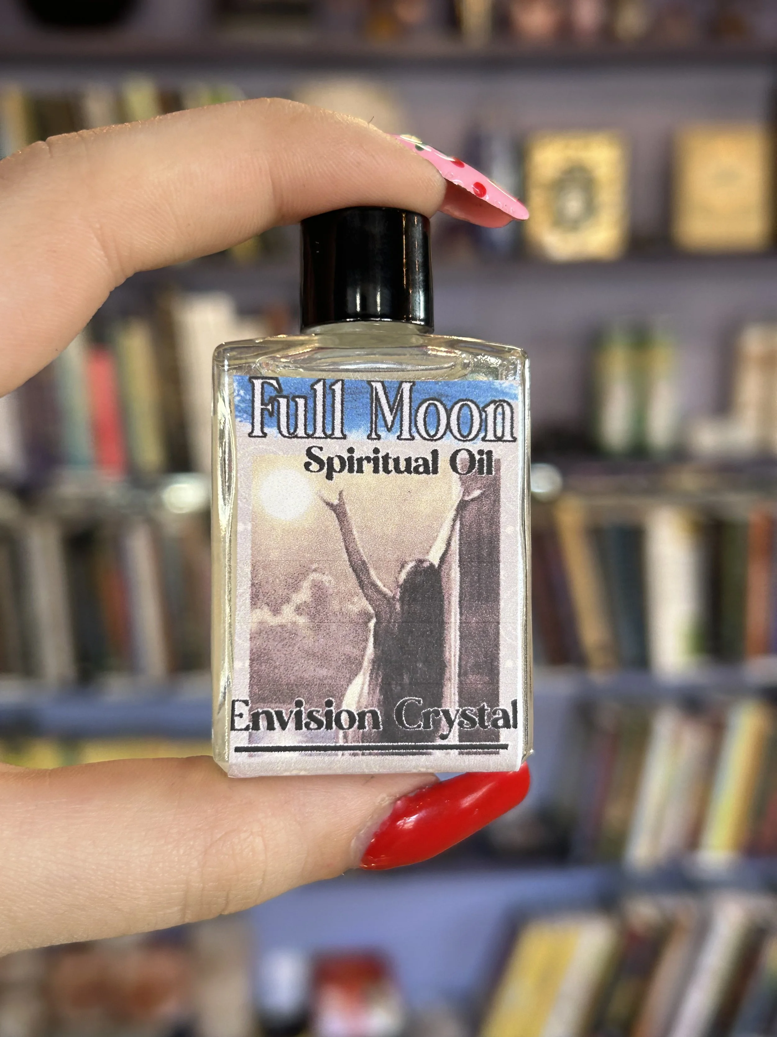 A square bottle is held in front of a purple bookshelf. It is labeled Full Moon Spiritual Oil with a classical drawing of a woman drawing down the energy of the moon.