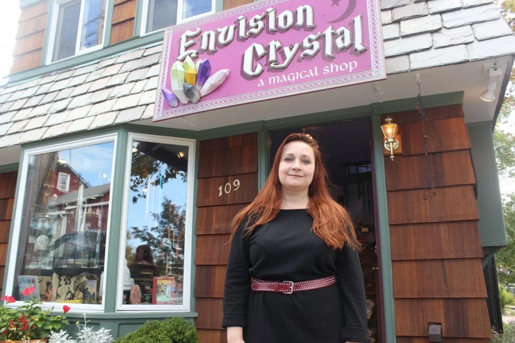 A redheaded woman named Lisa Mcgarrity is wearing a black dress, standing in front of her 'Envision Crystal' metaphysical shop with a colorful sign and wooden exterior.