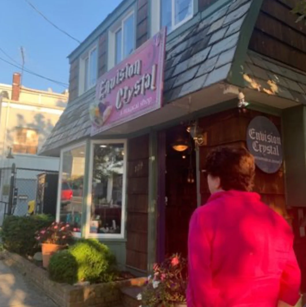 A person in a pink jacket approaching Envision Crystal, a store with a pink sign, outdoor plants, and a wooden facade.