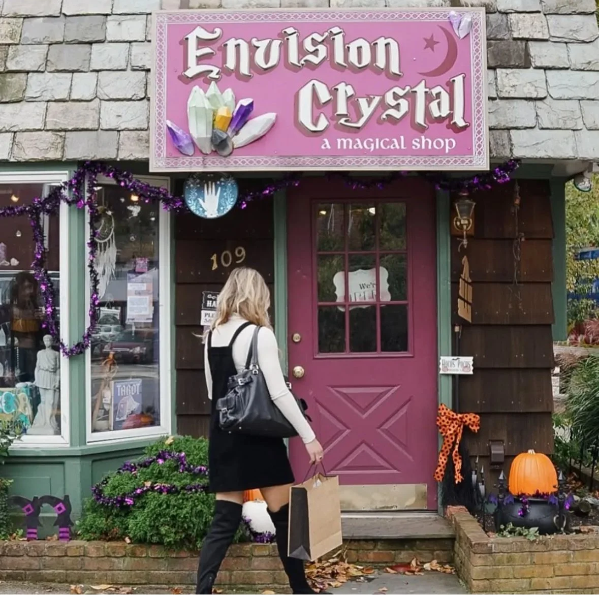 A woman carrying shopping bags approaches the entrance of a shop called 'Envision Crystal', which is described as a magical shop. The storefront is decorated with purple garlands and has Halloween-themed decorations, including pumpkins and autumn lea