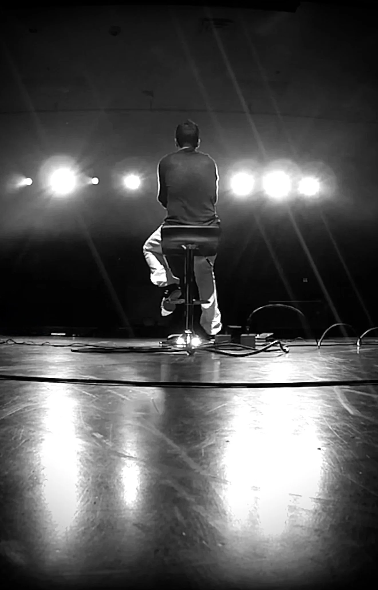Un homme assis sur un tabouret sur scène, vu de dos avec des lumières brillantes derrière lui, en noir et blanc.