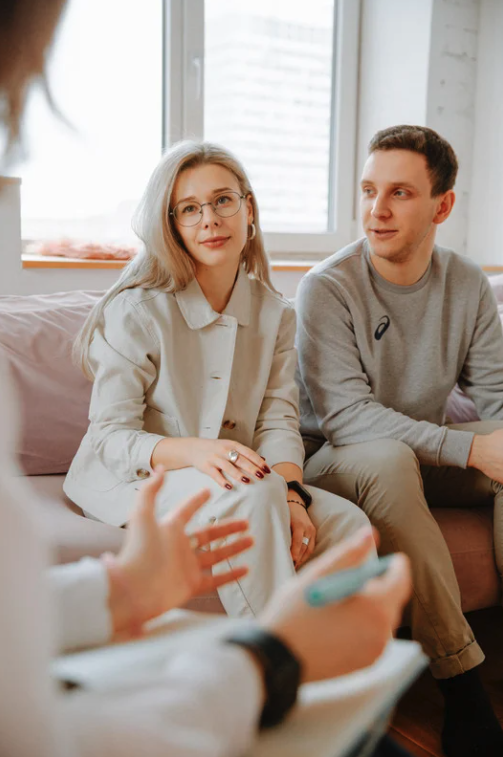 A woman with glasses and shoulder-length blonde hair sitting on a couch with a man beside her, both listening to a person speaking in a therapy or counseling session.