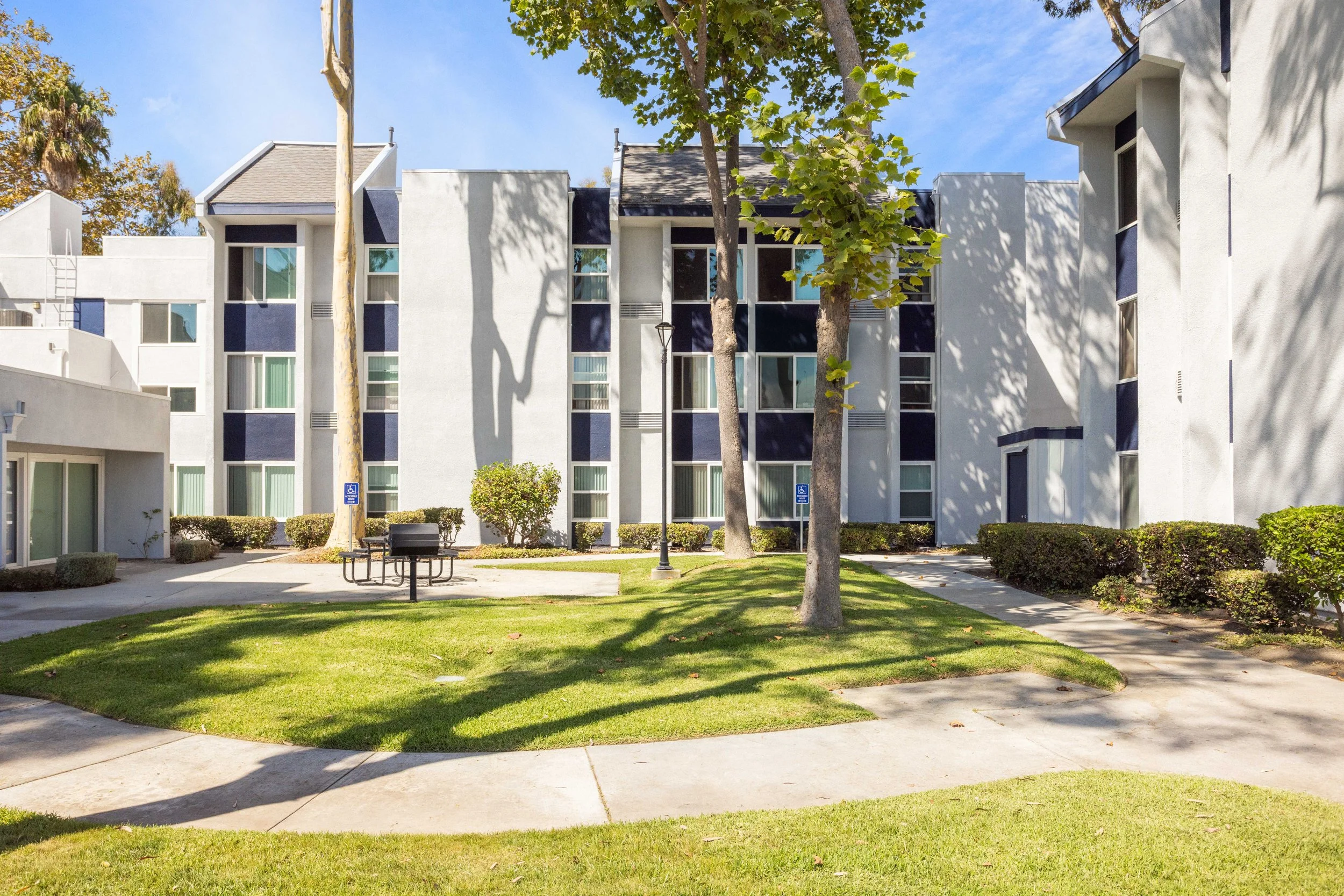 Exterior view of a modern white apartment building with multiple windows, surrounded by green grass, trees, and a sidewalk, under a blue sky.