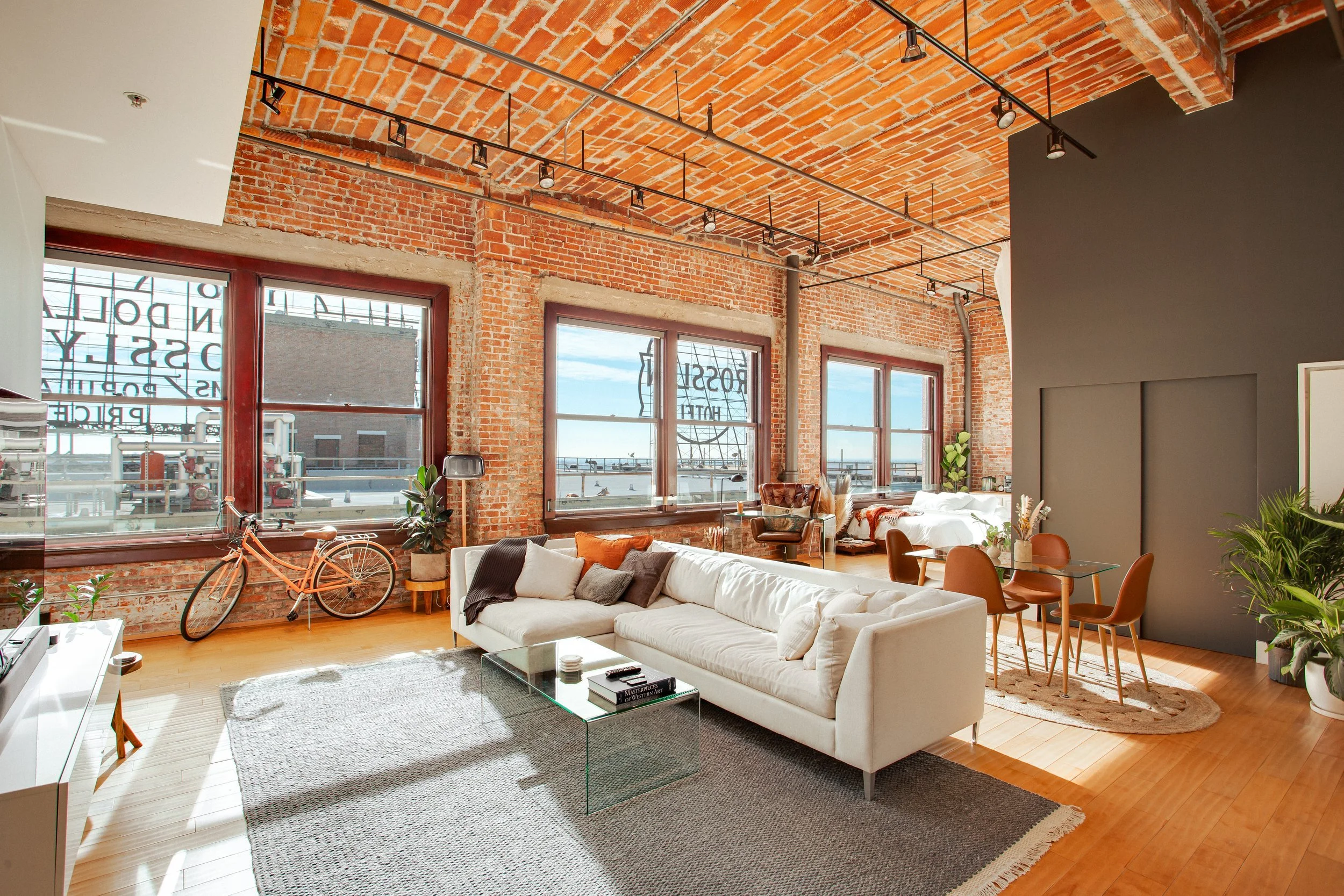 Modern living room with exposed brick walls, large windows, wooden floors, and a white sofa. Decor includes a glass coffee table, a bicycle by the window, indoor plants, a round rug, and a dining area with a table and chairs.
