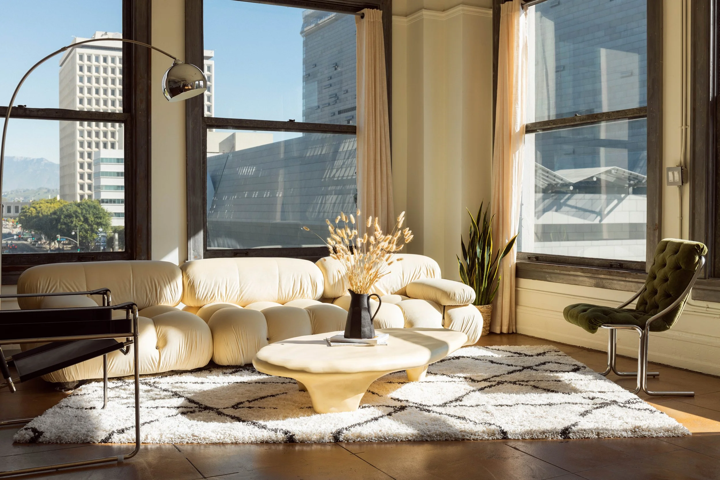 Living room with large windows, cream-colored cushioned sofa, green armchair, coffee table with vase of dried flowers, and potted plants, flooded with sunlight.