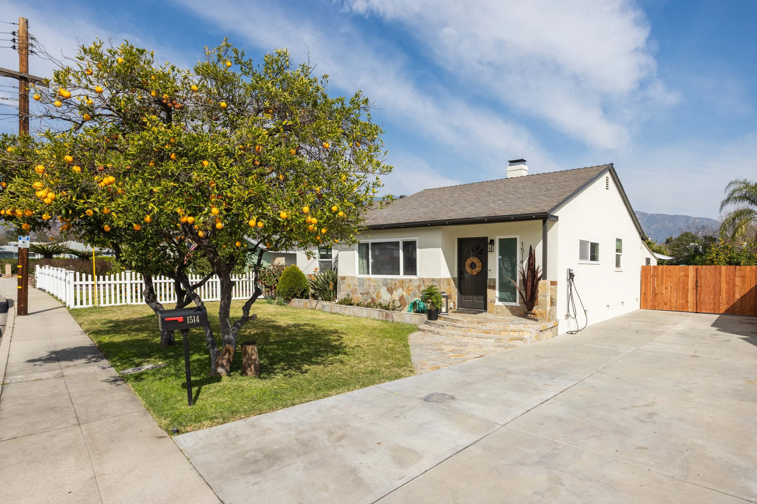 White house with stone accents on the front, a tree with orange fruit, a mailbox, and a paved driveway. A white picket fence is on the left, and a wooden fence is on the right. The sky is partly cloudy.