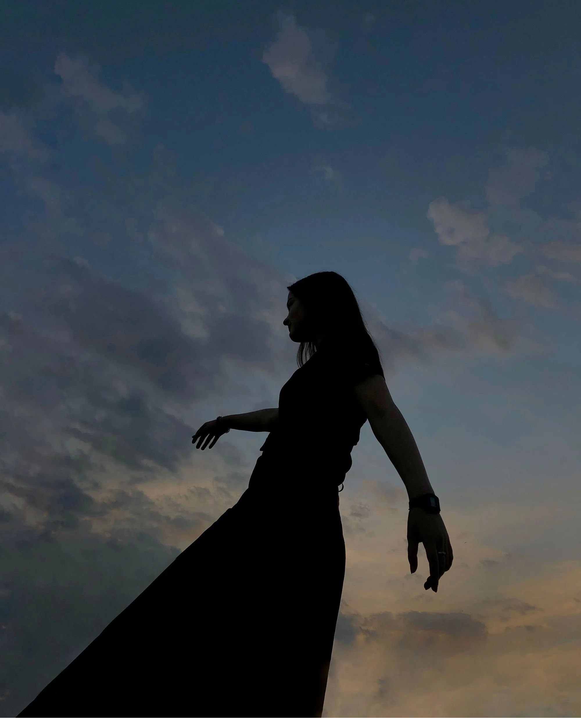 Silhouette of a woman with long hair standing against a sky at sunset or dusk, with clouds and a gradient of dark to light colors.
