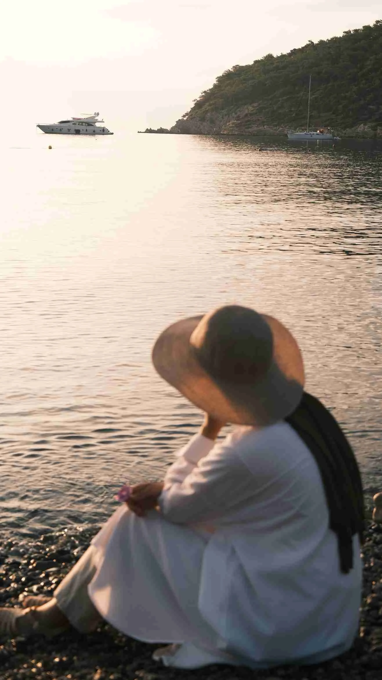 Person wearing a white outfit and a wide-brimmed hat sitting on a rocky shoreline, looking out at the water with boats and a hillside in the distance during sunset.