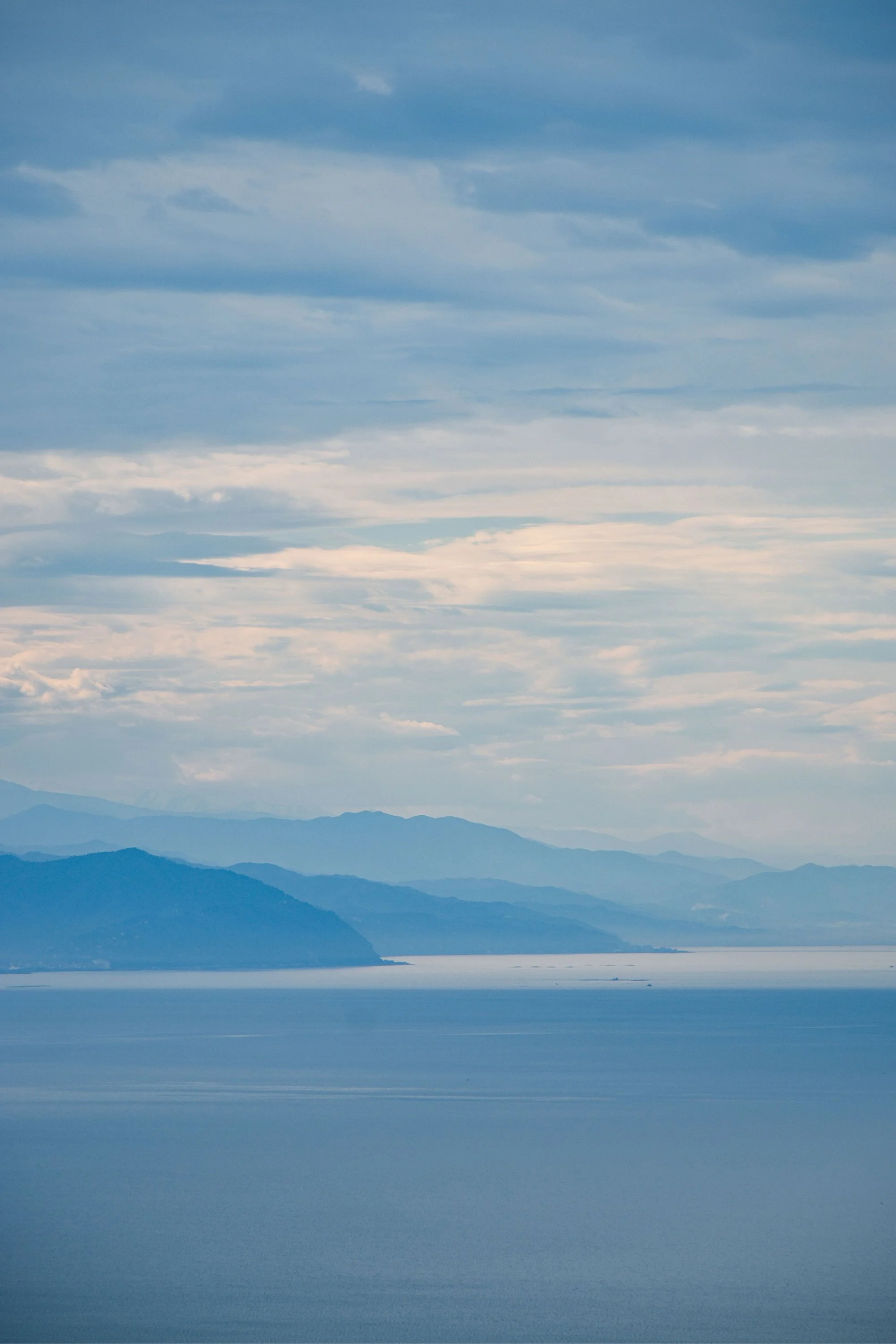 A serene landscape with multiple layers of distant blue mountains under a cloudy sky over a calm body of water.