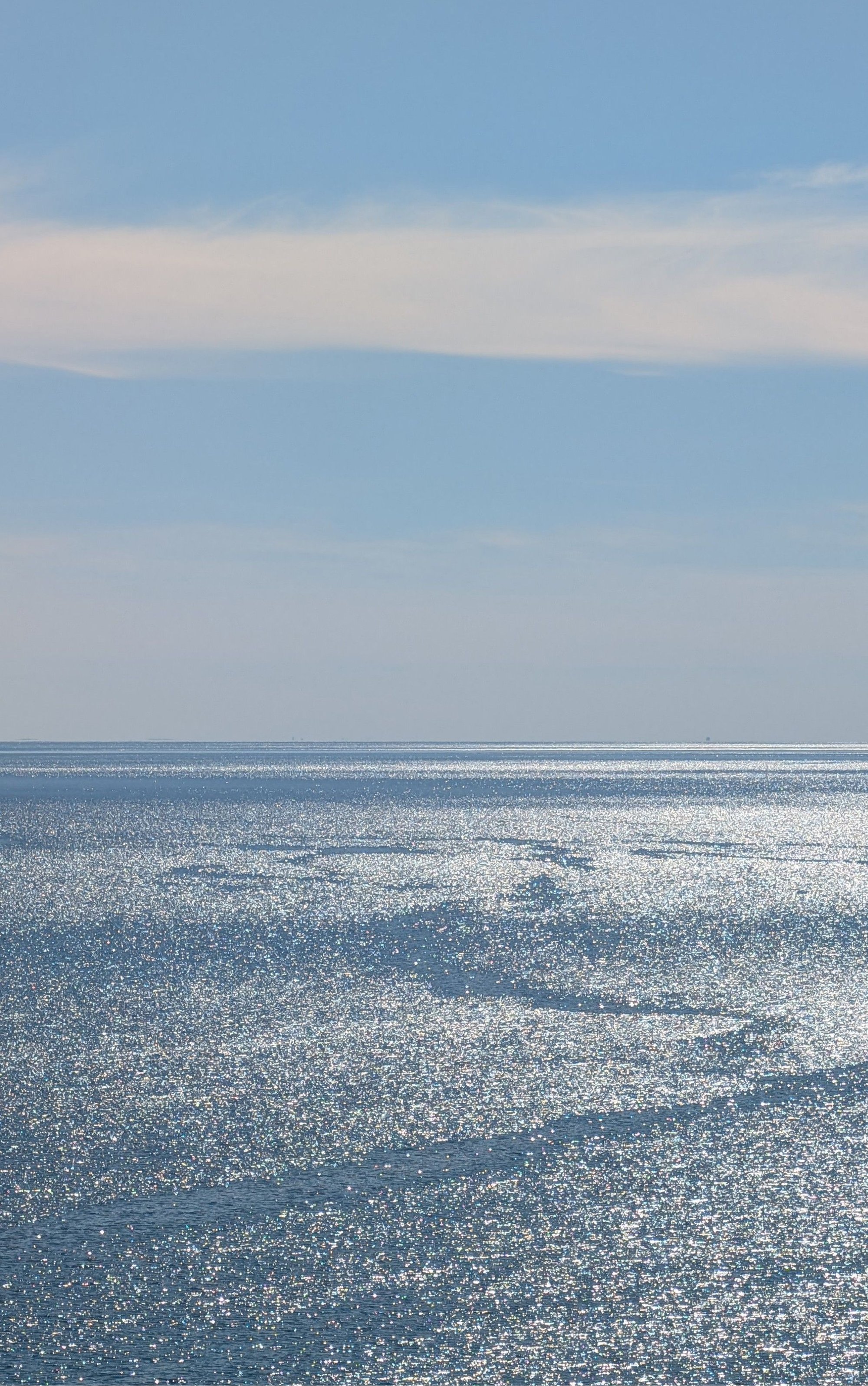 Shimmering ocean with sunlight reflecting off the water, under a clear blue sky with some wispy clouds.