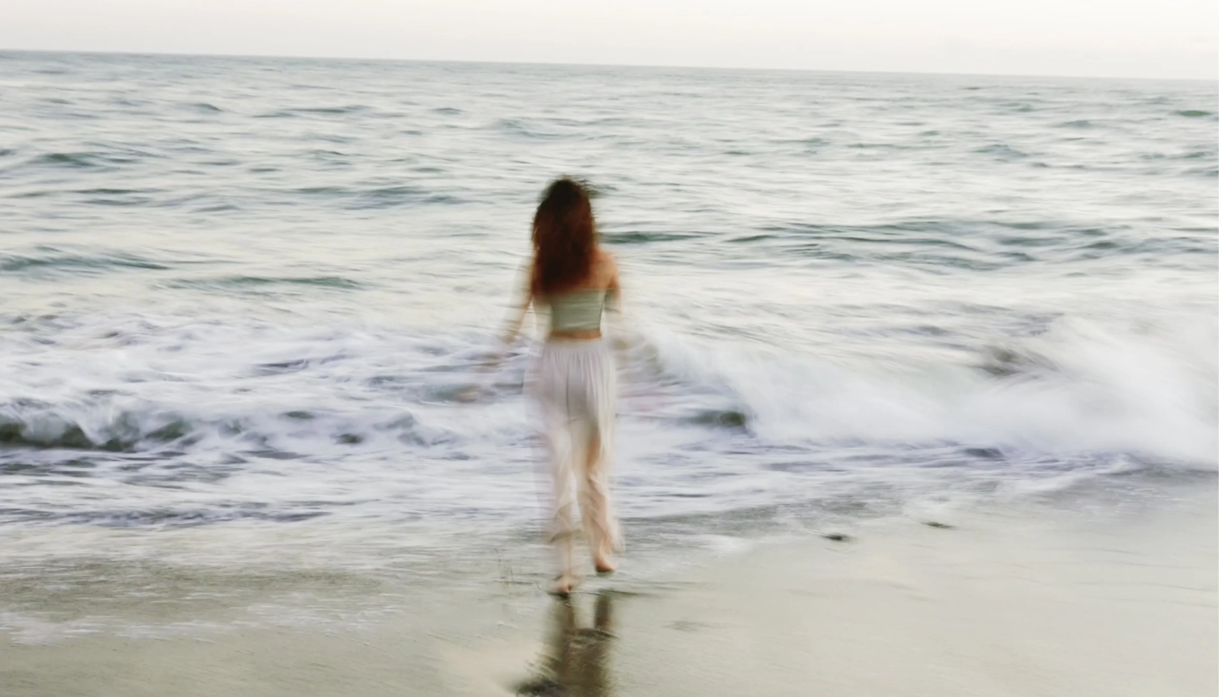 A woman with long hair, wearing a strapless top and flowing pants, walking into the ocean waves on a sandy beach, viewed from behind.